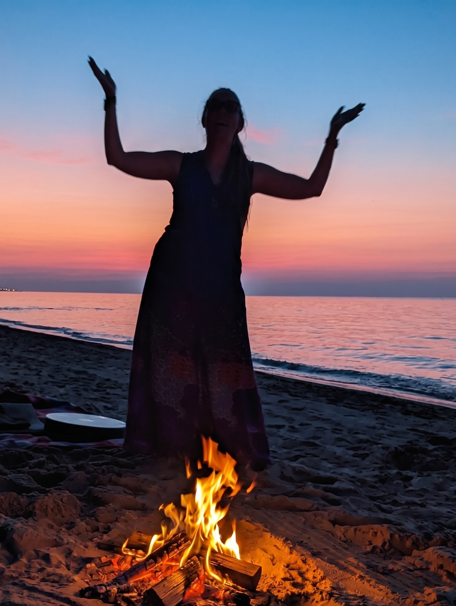 Person standing on a beach with arms raised, wearing glasses, near a campfire at sunset, with water and colorful sky in the background.