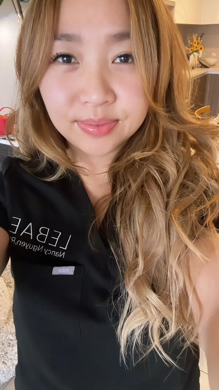 Close-up of a woman with long, wavy, blonde hair and light skin, smiling with natural makeup, in a room with a beige background, a counter, and decorative items.