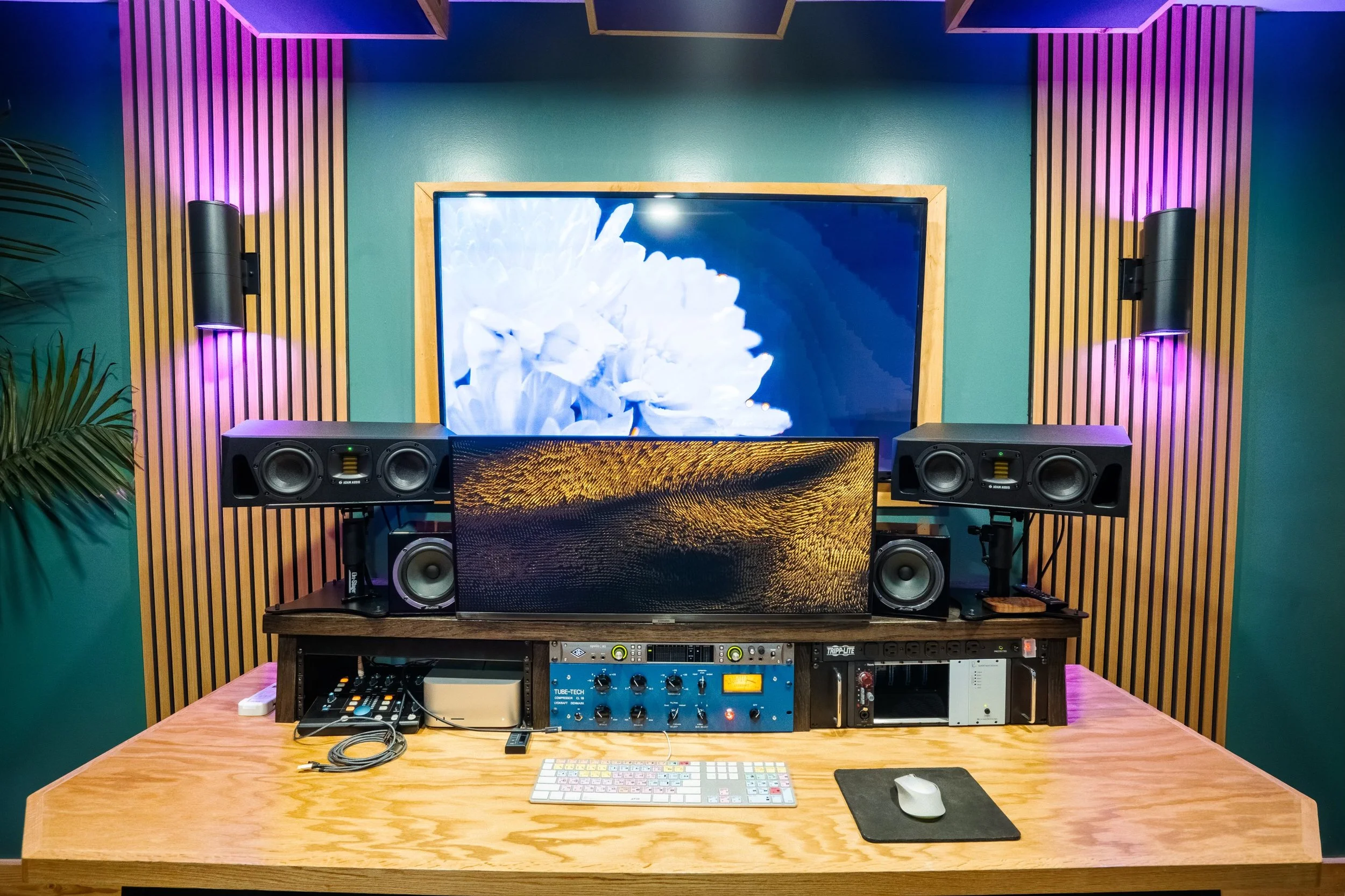 Modern audio mixing studio with multiple monitors, speakers, and sound equipment on a wooden desk in a room with teal walls and acoustic panels.