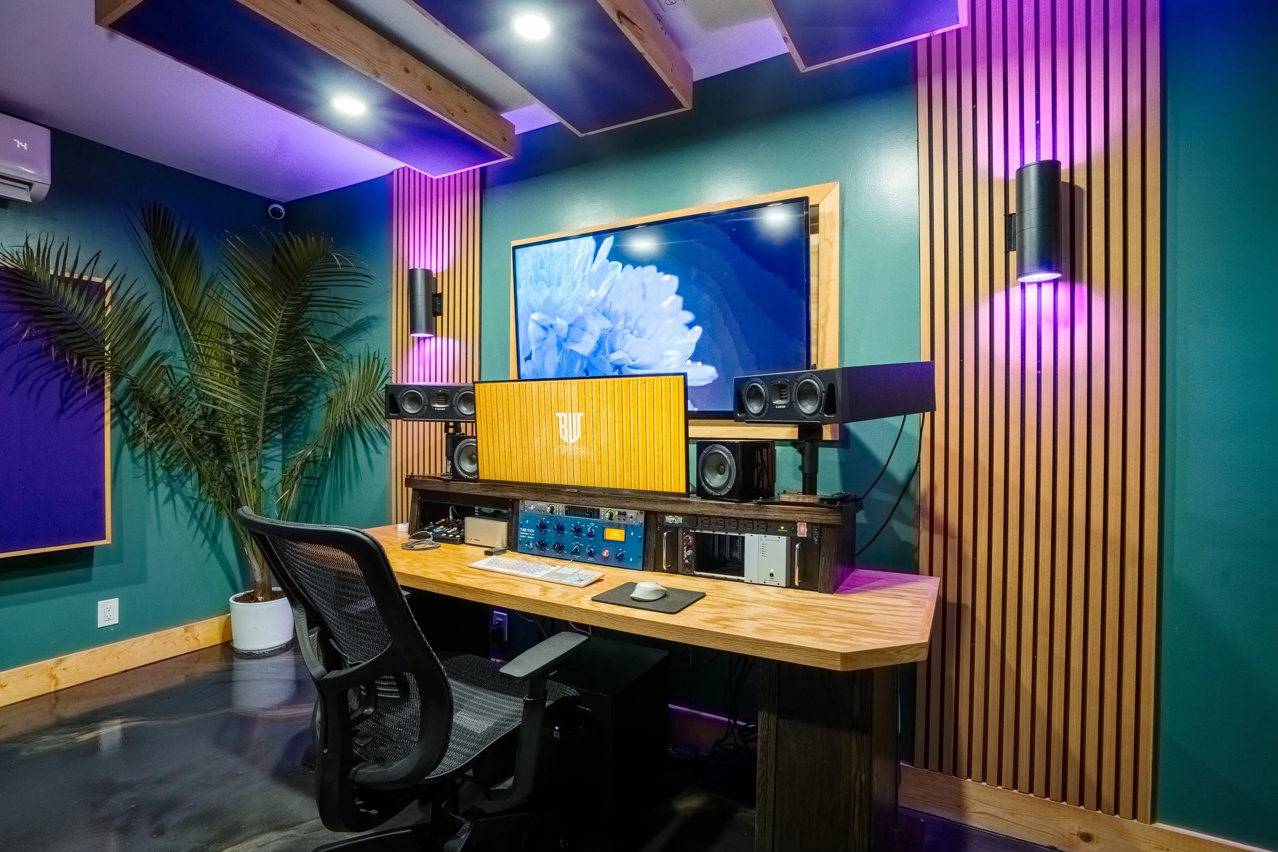Modern recording studio with a desk, speakers, and acoustic panels, featuring purple ambient lighting and a large monitor displaying a flower image.