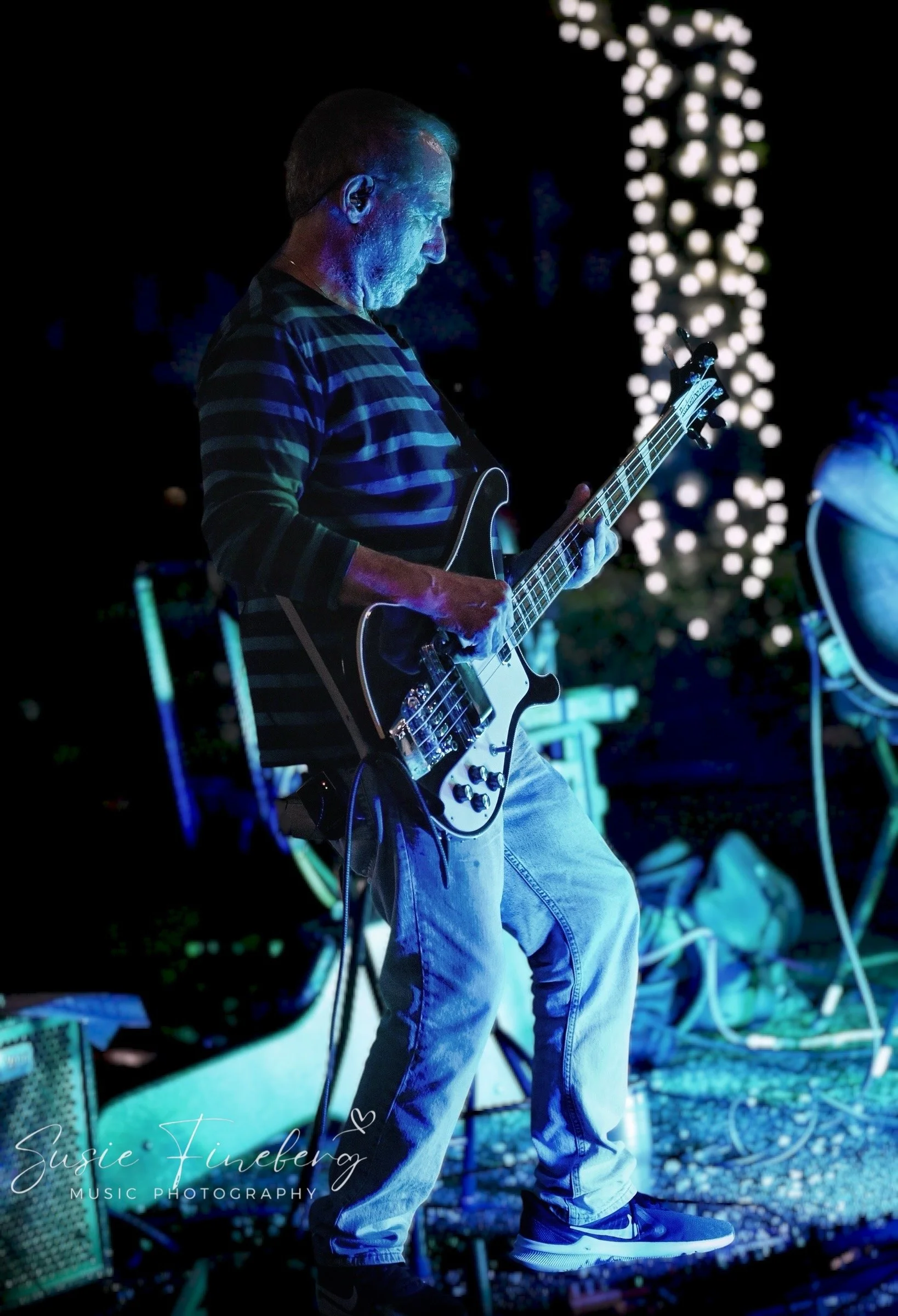 Man playing electric guitar during a nighttime outdoor performance with backdrop of decorative illuminated trees.