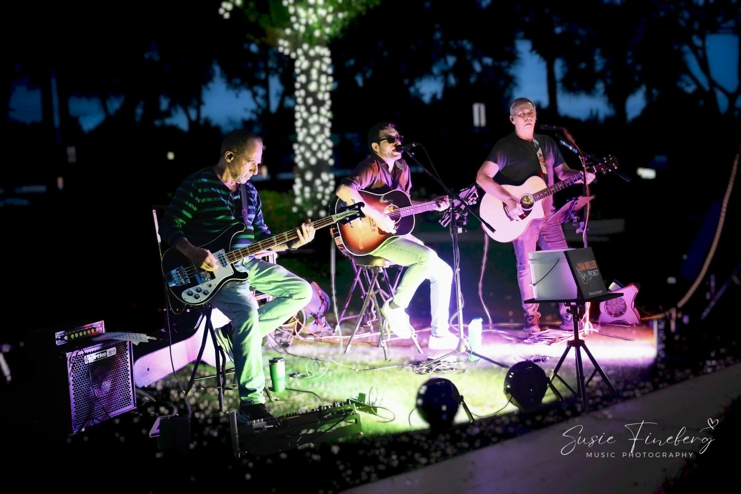 Three musicians performing at night with a dark sky and illuminated tree in the background. They are sitting and standing, playing guitars and singing into microphones, under colorful stage lighting.