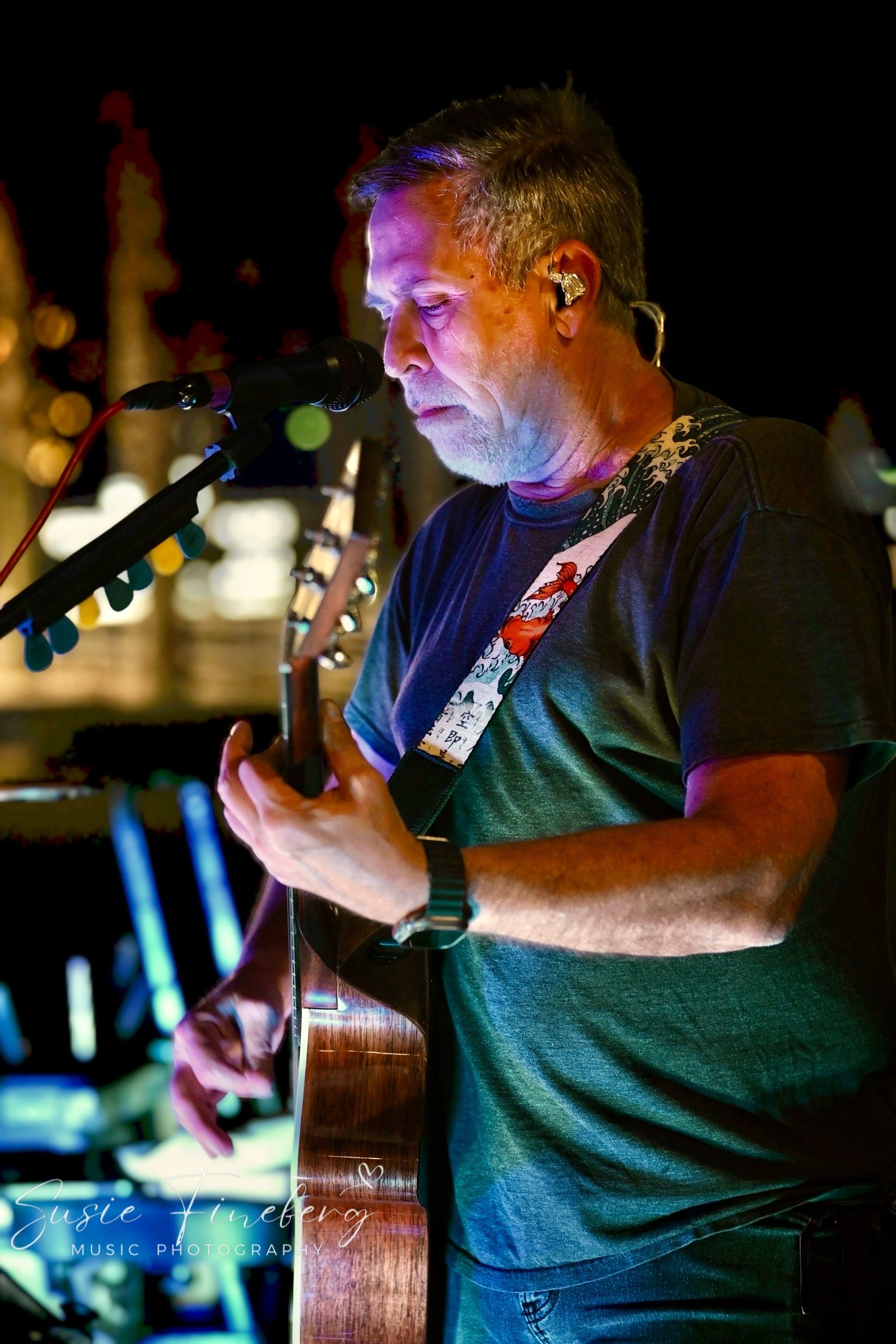 Man performing live music on stage at night, playing an acoustic guitar and singing into a microphone, with blurred colorful background lights.