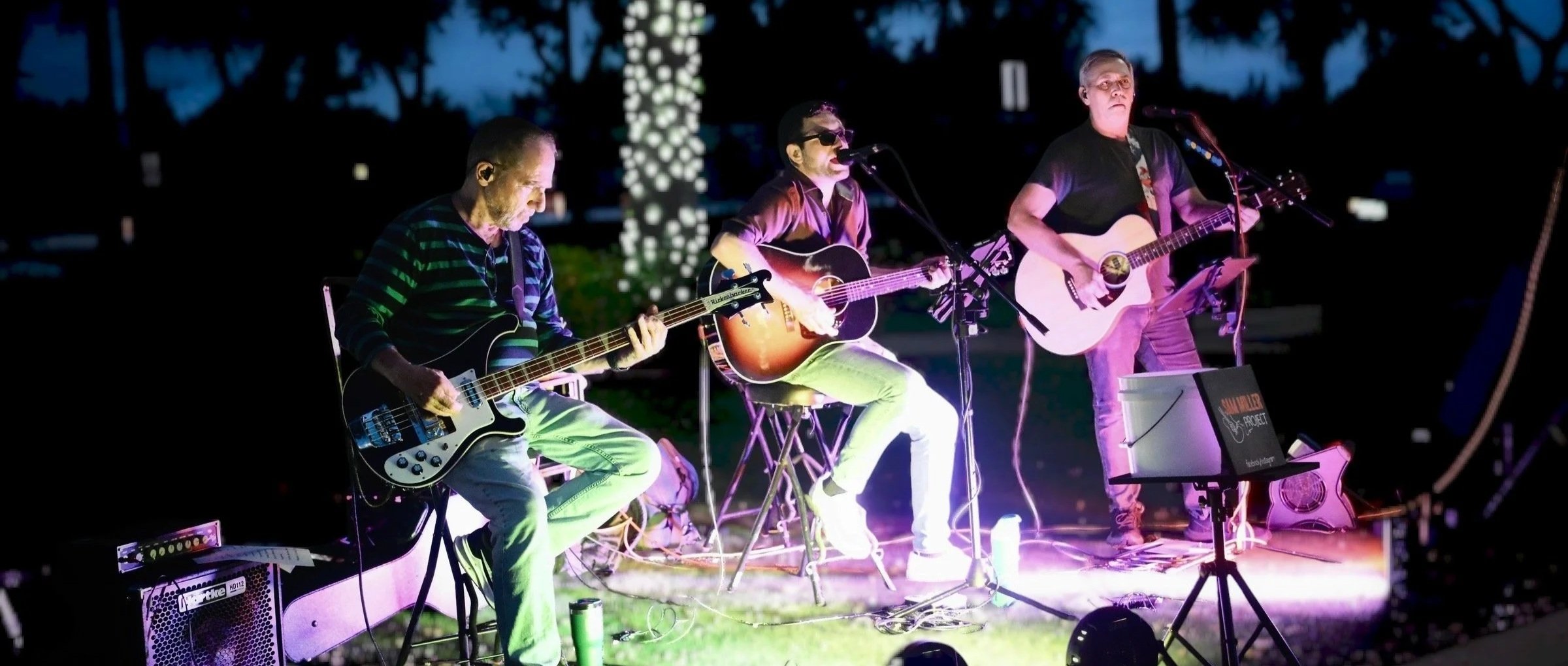 Three musicians performing with guitars on an outdoor stage at night, illuminated by colorful stage lights, with dark trees in the background.