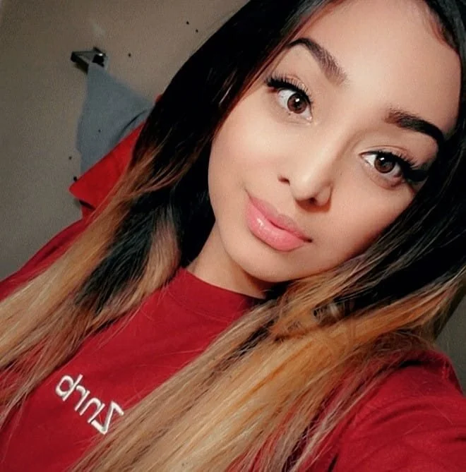 A young woman with long, wavy hair and light makeup taking a selfie indoors. She is wearing a red shirt.