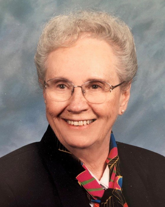 Portrait of an elderly woman with short gray hair, wearing glasses, a dark blazer, and a colorful scarf, smiling against a blue background.