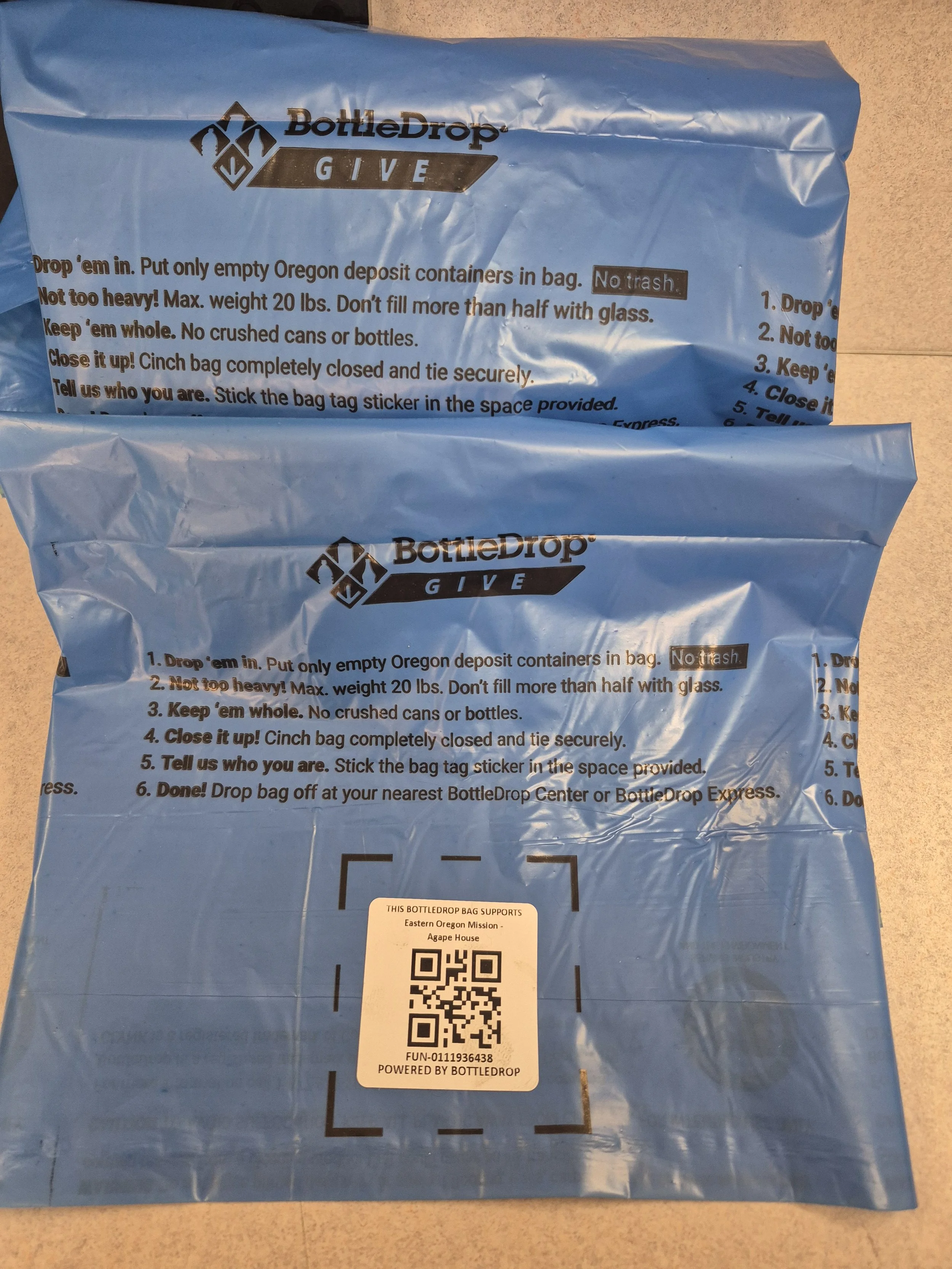Two blue BottleDrop GIVE bags with instructions and a QR code printed on them, placed on a light-colored surface.