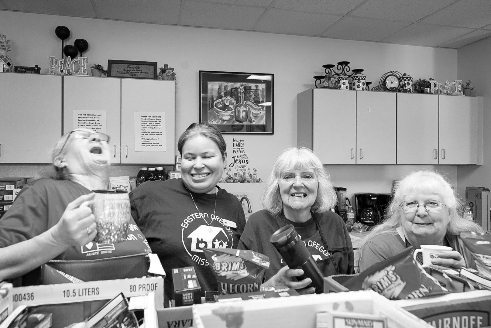 Four women smiling and laughing around a table filled with food and drinks, in a kitchen. The woman on the left is holding a large mug, the second woman is wearing a dark shirt, the third woman with gray hair is holding a flashlight, and the fourth woman is drinking from a mug. Kitchen cabinets and decorations are in the background.
