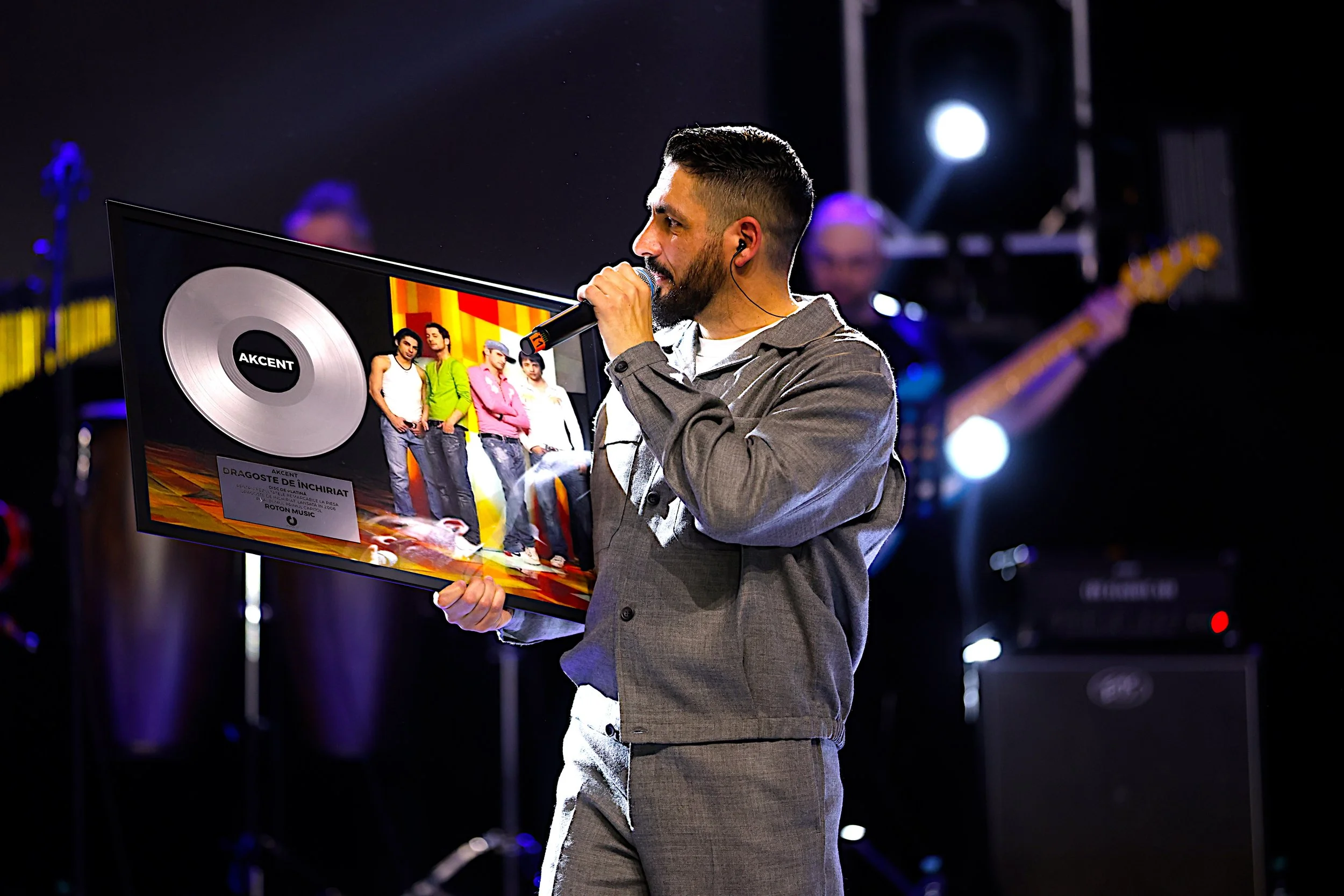 A man with a beard and short dark hair, wearing a gray suit, speaks into a microphone while holding a framed picture of a music album cover on stage. The album cover features five young people standing together in casual clothing. The stage is lit with bright lights and a band is visible in the background.