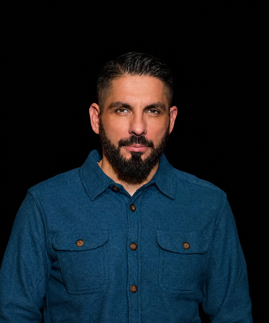 A man with dark hair and a beard, wearing a blue button-up shirt, posing against a solid black background.