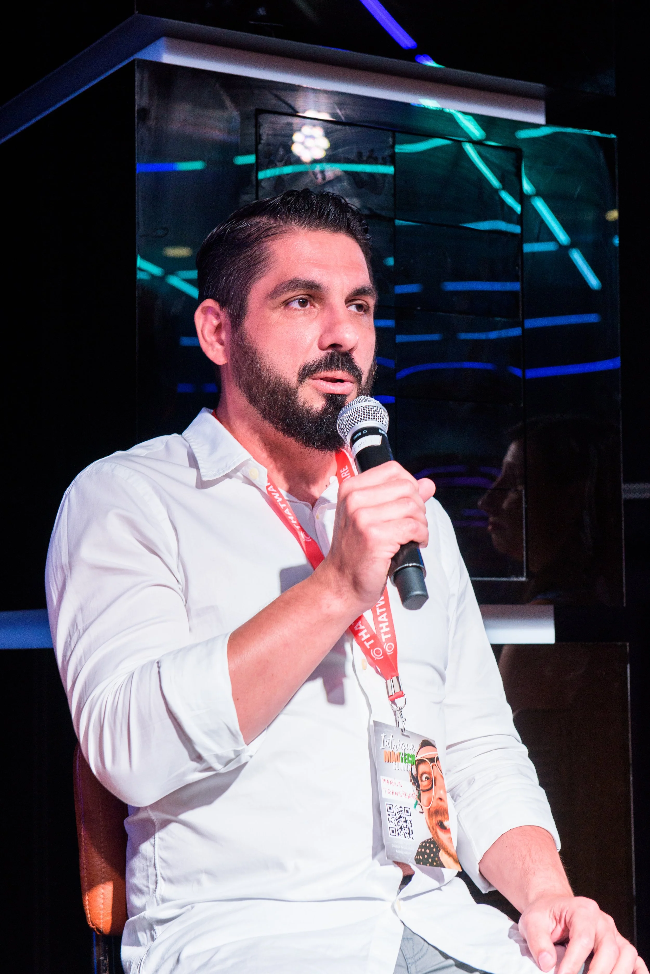 A man with dark hair, a beard, and light skin is sitting and speaking into a microphone. He is wearing a white shirt and a lanyard with a badge that has a cartoon image and QR code. Behind him is a reflective, dark surface with colorful neon lights.