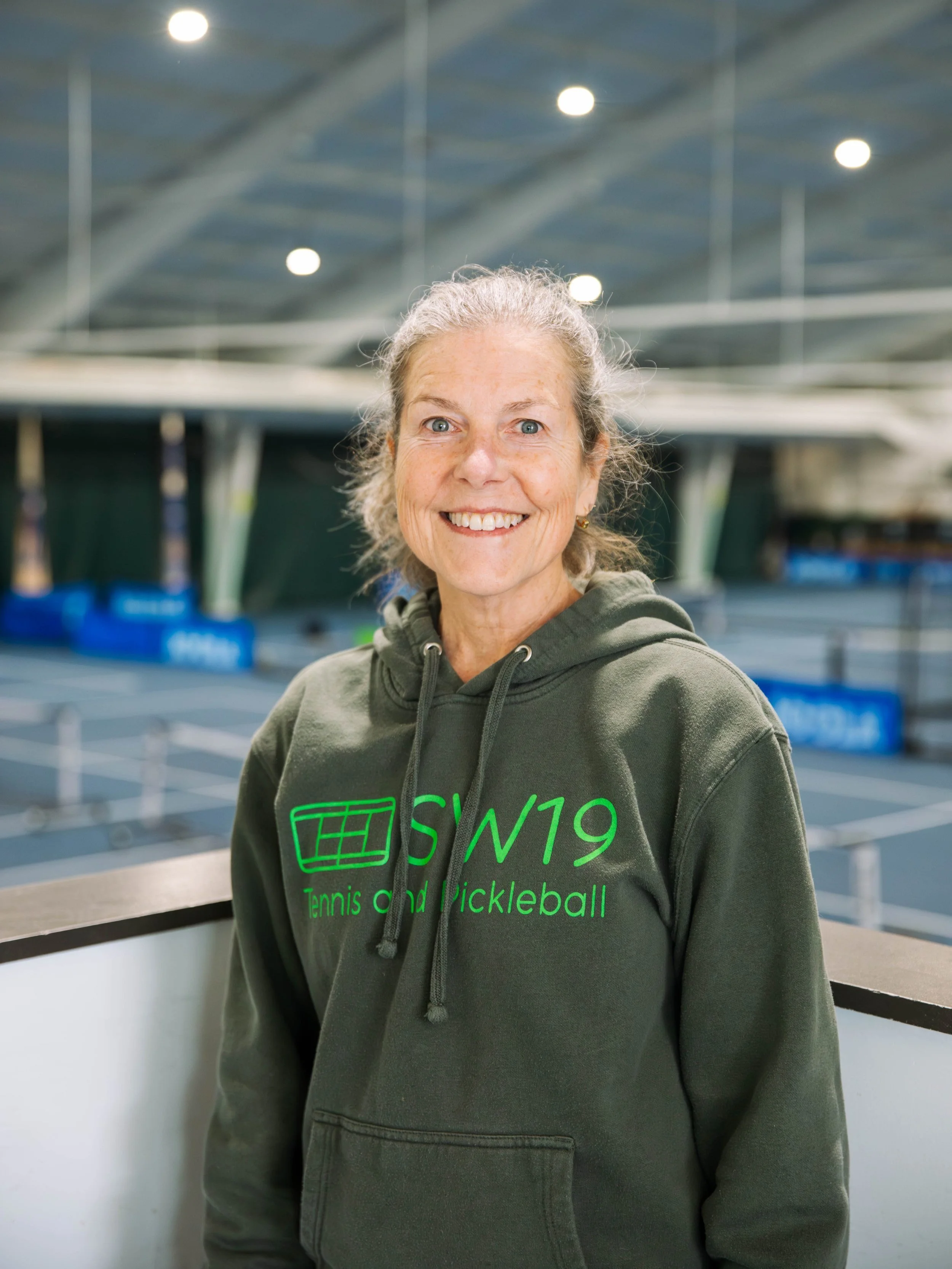 A person wearing a green hoodie with 'SW19 Tennis and Pickleball' in a sports facility.
