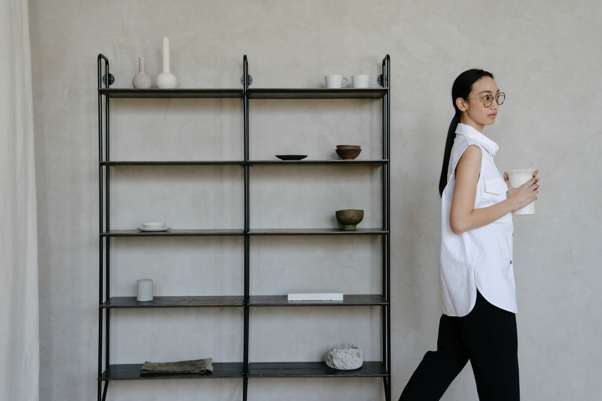 Woman with glasses, long black hair, wearing a white sleeveless vest and black pants, holding a white mug, walking past a black metal open shelf with various ceramic and stone objects against a plain beige wall.