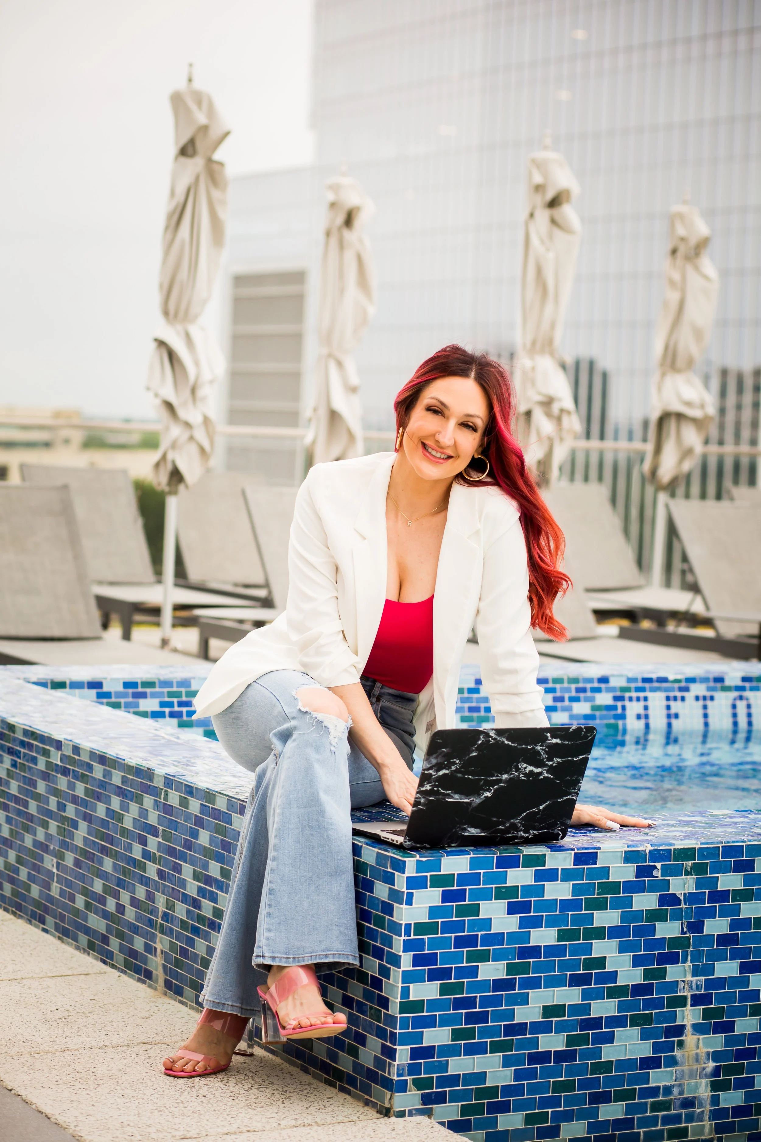 A woman with red hair and a white blazer sitting on the edge of a small pool with a laptop, smiling at the camera, with poolside lounge chairs and umbrellas in the background.