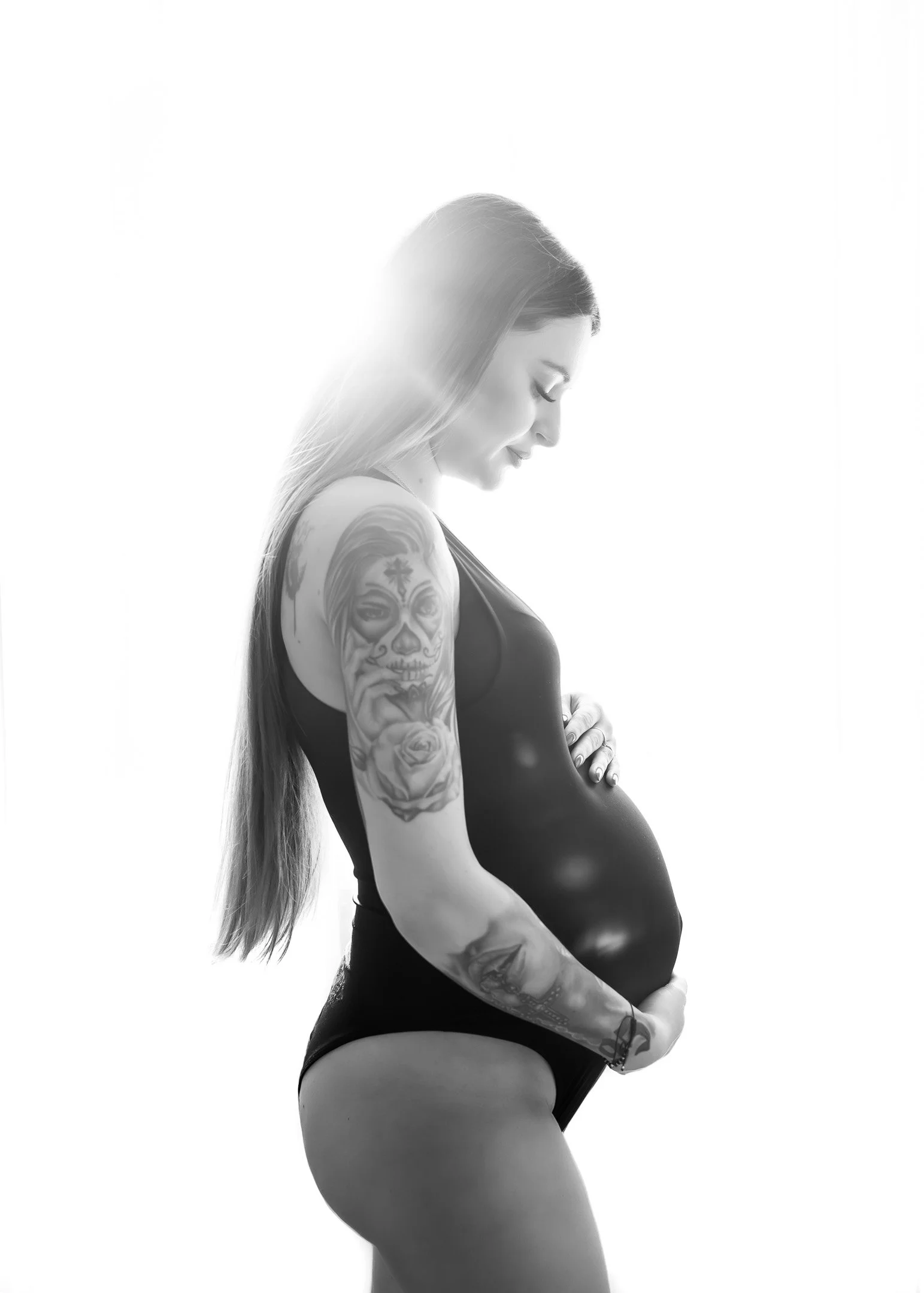 Black and white photo of pregnant woman with long hair, tattoos on arm, wearing black underwear, standing sideways with eyes closed and hand on her belly.