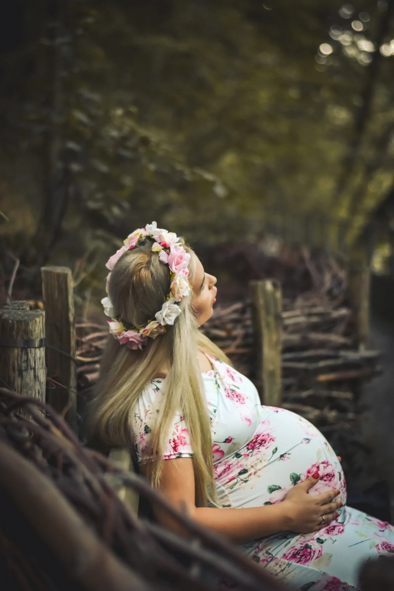 Pregnant woman with floral headband sitting on a wooden bench in a wooded area, gently touching her belly.
