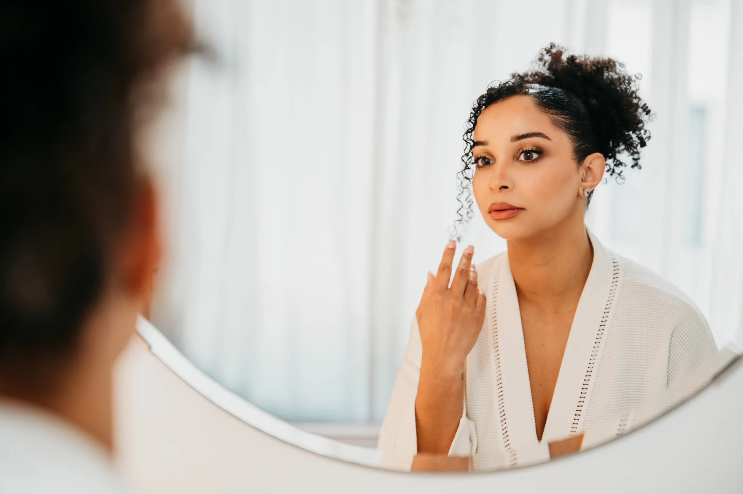 Woman looking at her reflection in a mirror, touching her face, in a well-lit room.