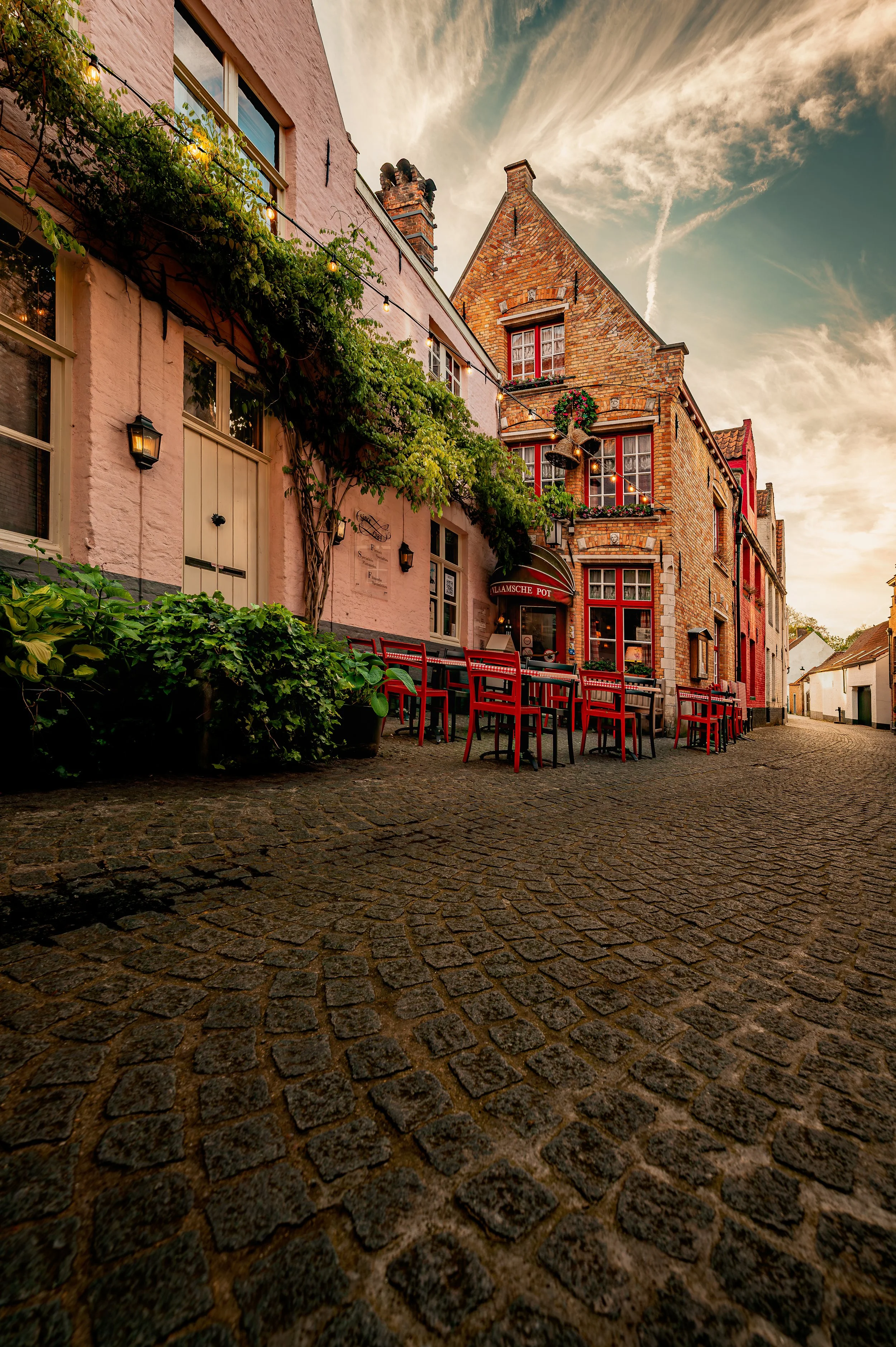 Outdoor cafe with red chairs and cobblestone street in a European village at sunset, with old brick and stucco buildings decorated with greenery and string lights.