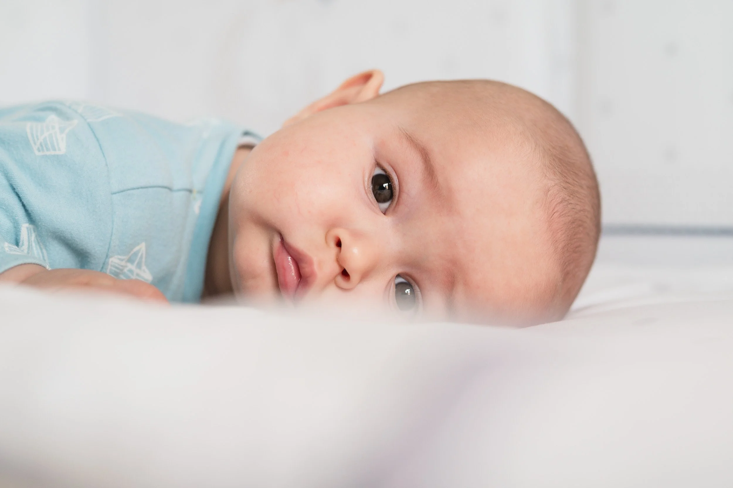 A close-up of a baby lying on its side, looking at the camera with big, brown eyes and a slight smile, dressed in a light blue onesie with white patterns.