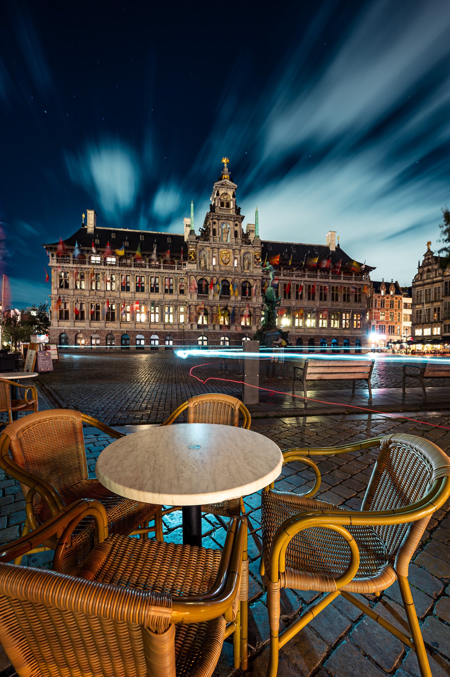 Empty outdoor cafe table with wicker chairs in front of illuminated historic building with a statue in a city square at night.
