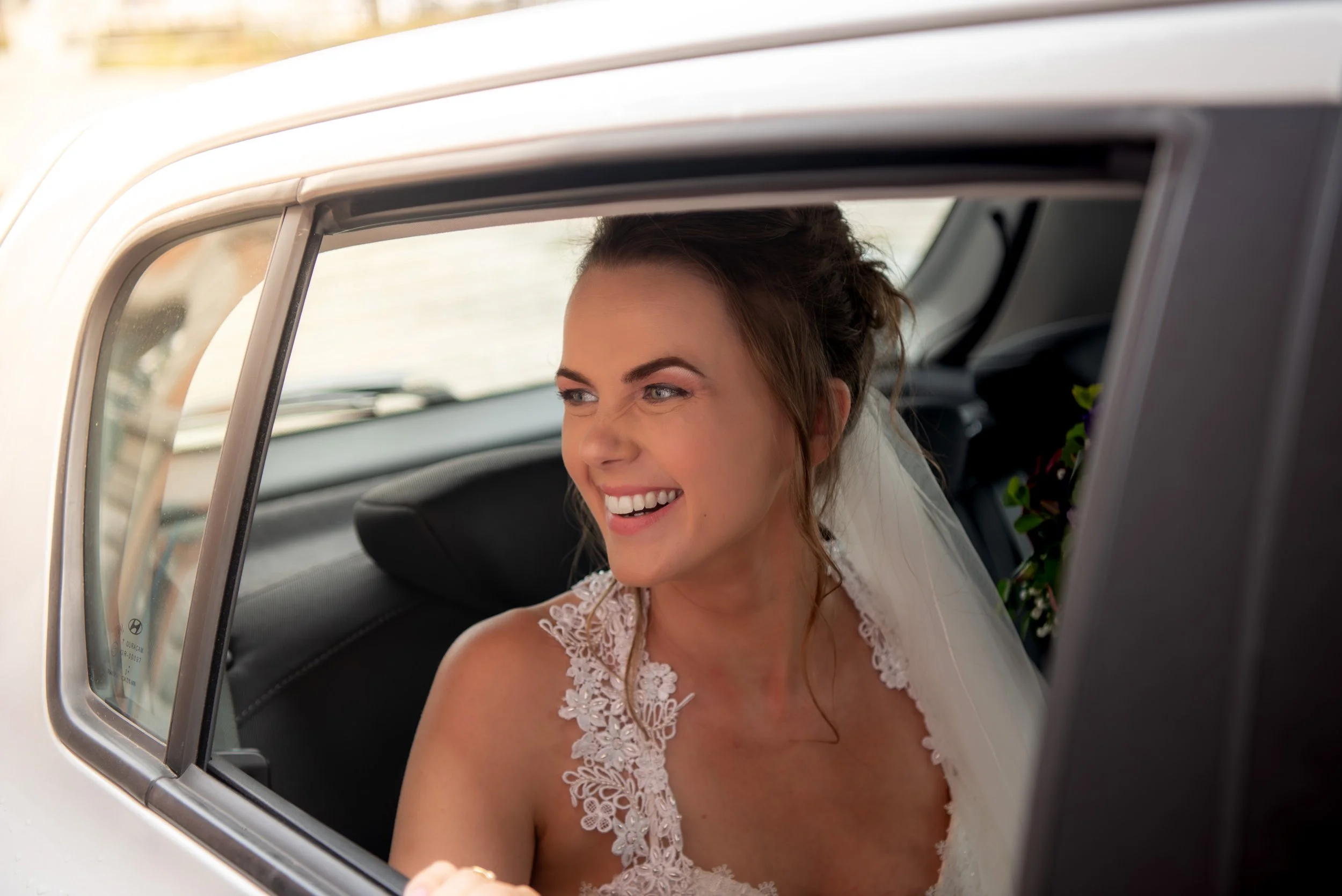 A smiling bride sitting in the backseat of a white car, wearing a lace wedding dress and veil, looking out the window.