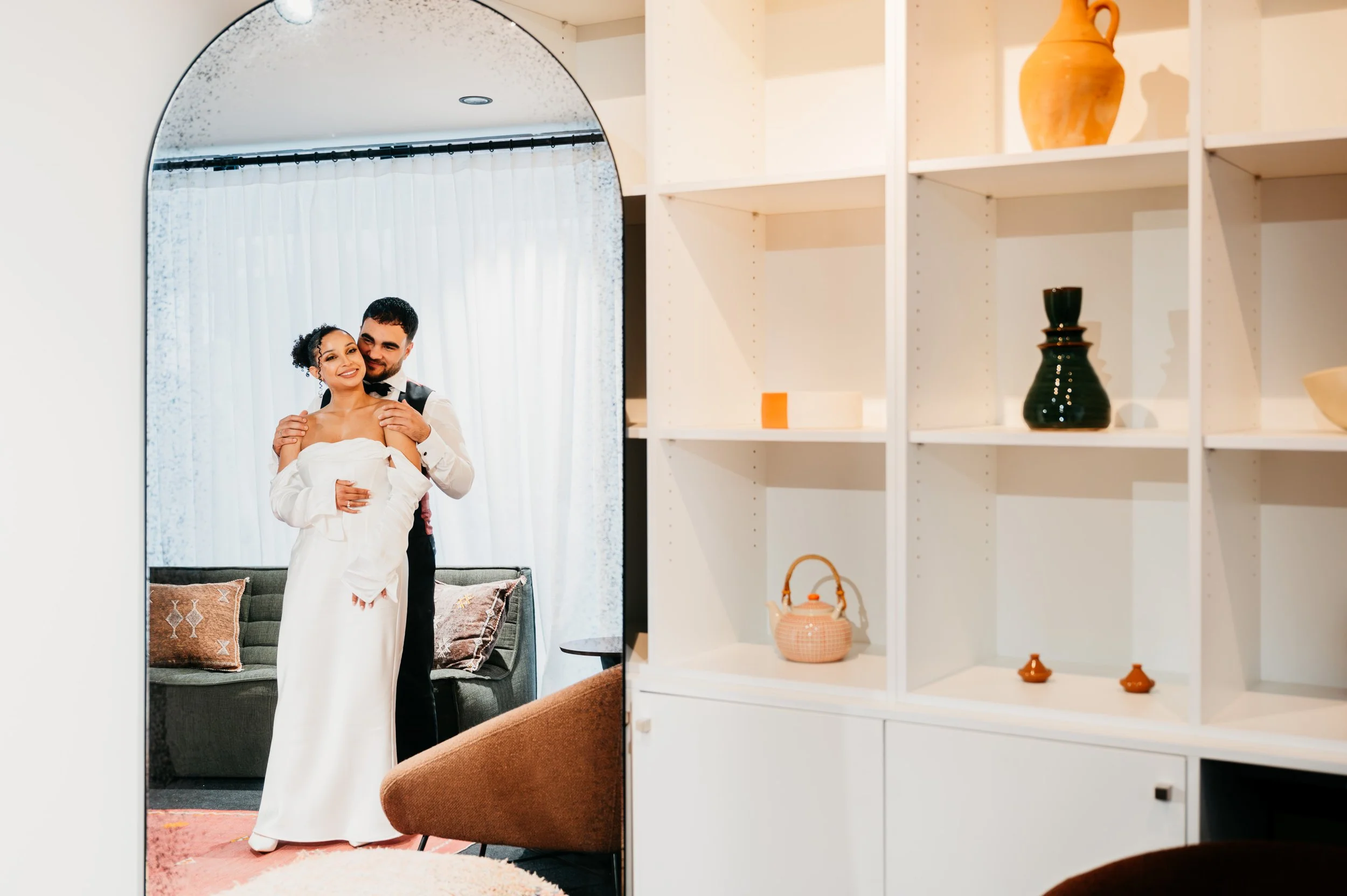 A bride, dressed in a white gown, smiles as a groom, in a suit, stands behind her holding her gently from behind in front of a mirror. They are in a cozy, well-decorated room with bookshelves and vases.