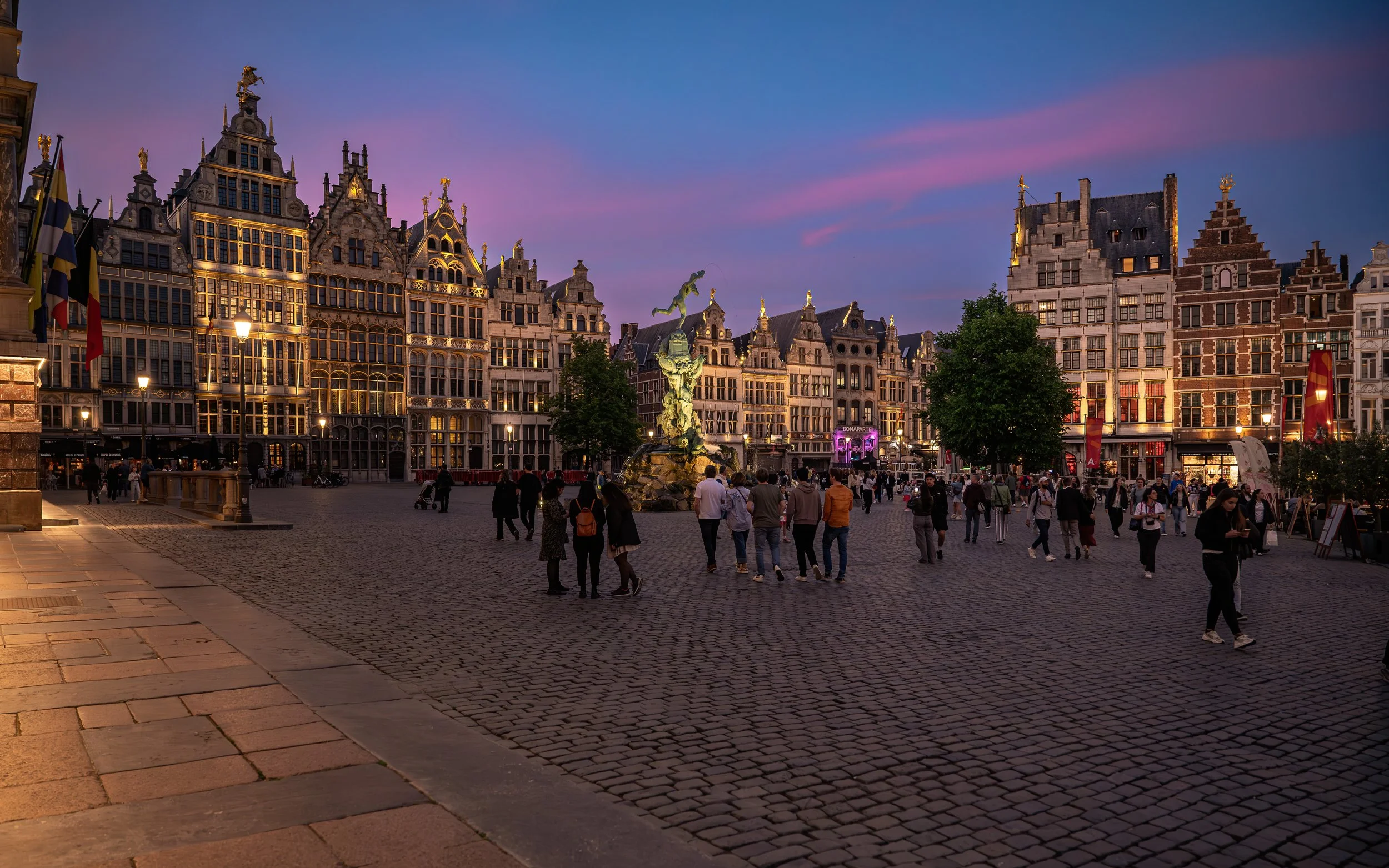 An evening scene at the Grand Place in Brussels, Belgium, with historic buildings, a fountain at the center, and people walking in the cobblestone square during sunset.