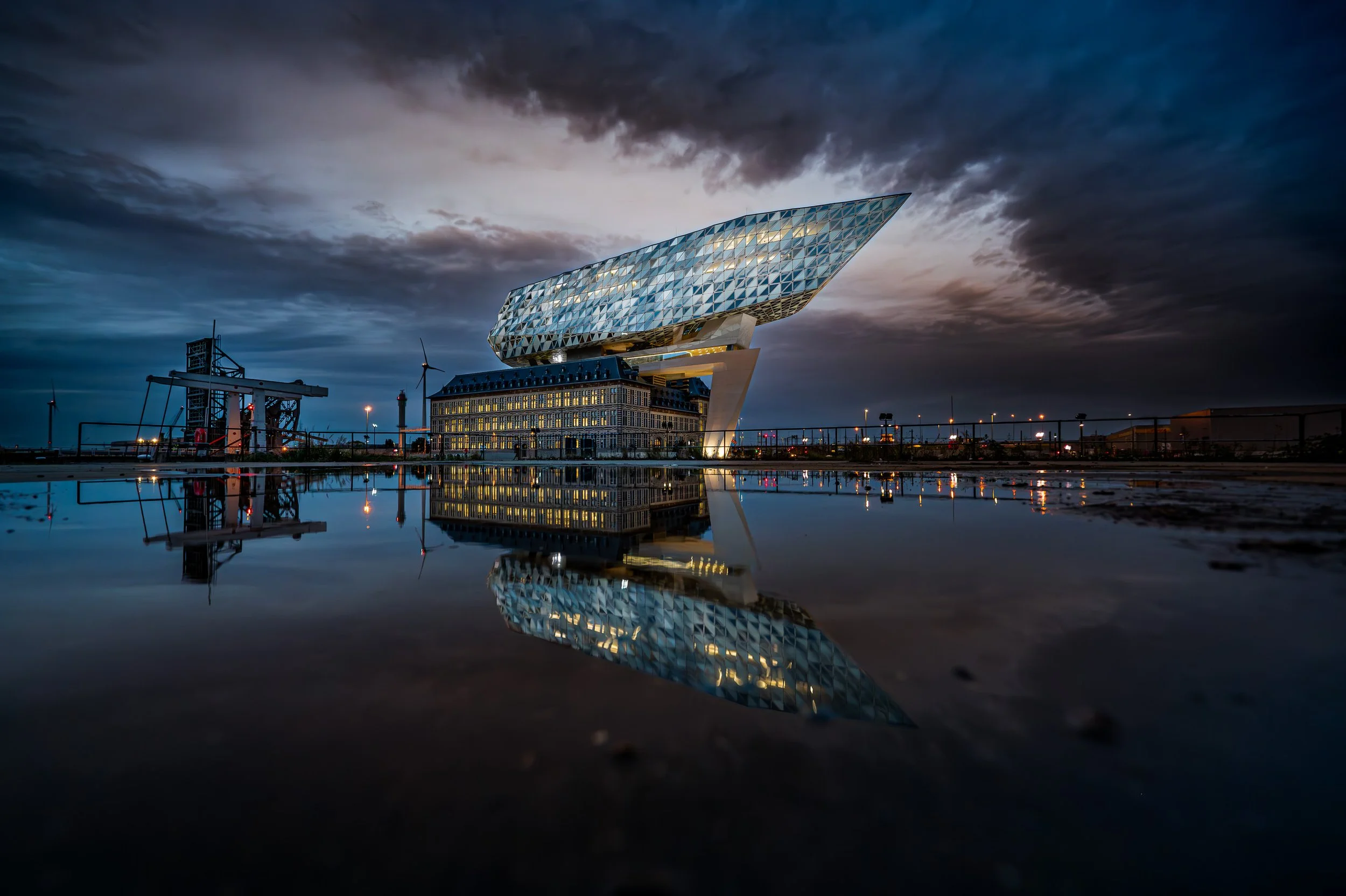Modern architectural building with an overhanging, diamond-patterned glass structure, reflected in a large pool of water at dusk under a cloudy sky.