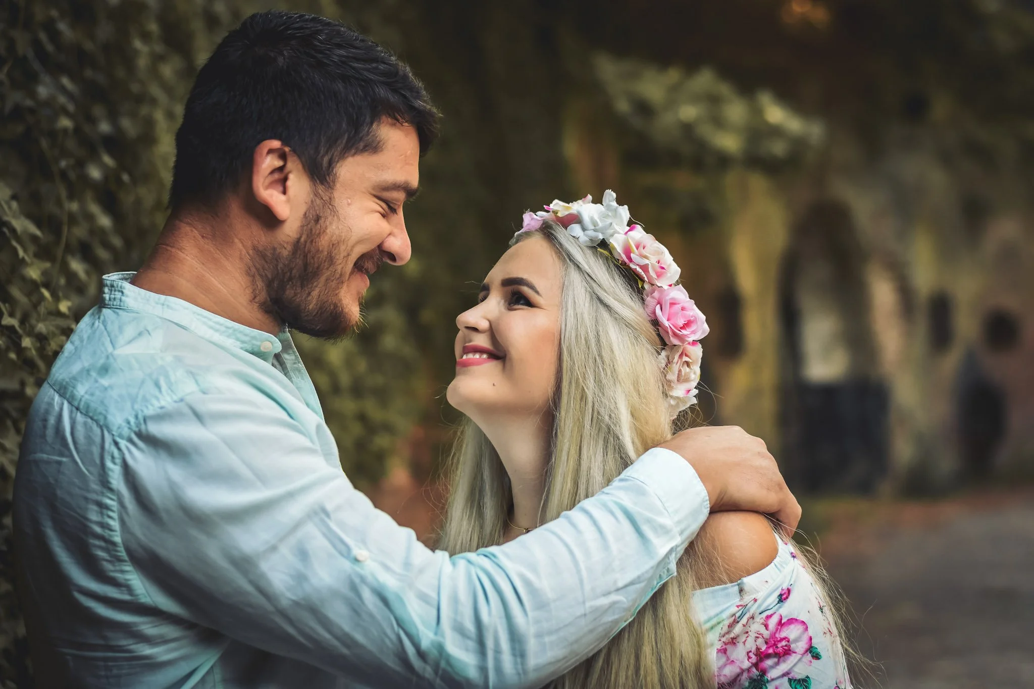 A couple smiling and looking at each other, with the man embracing the woman, outdoors in a wooded area.
