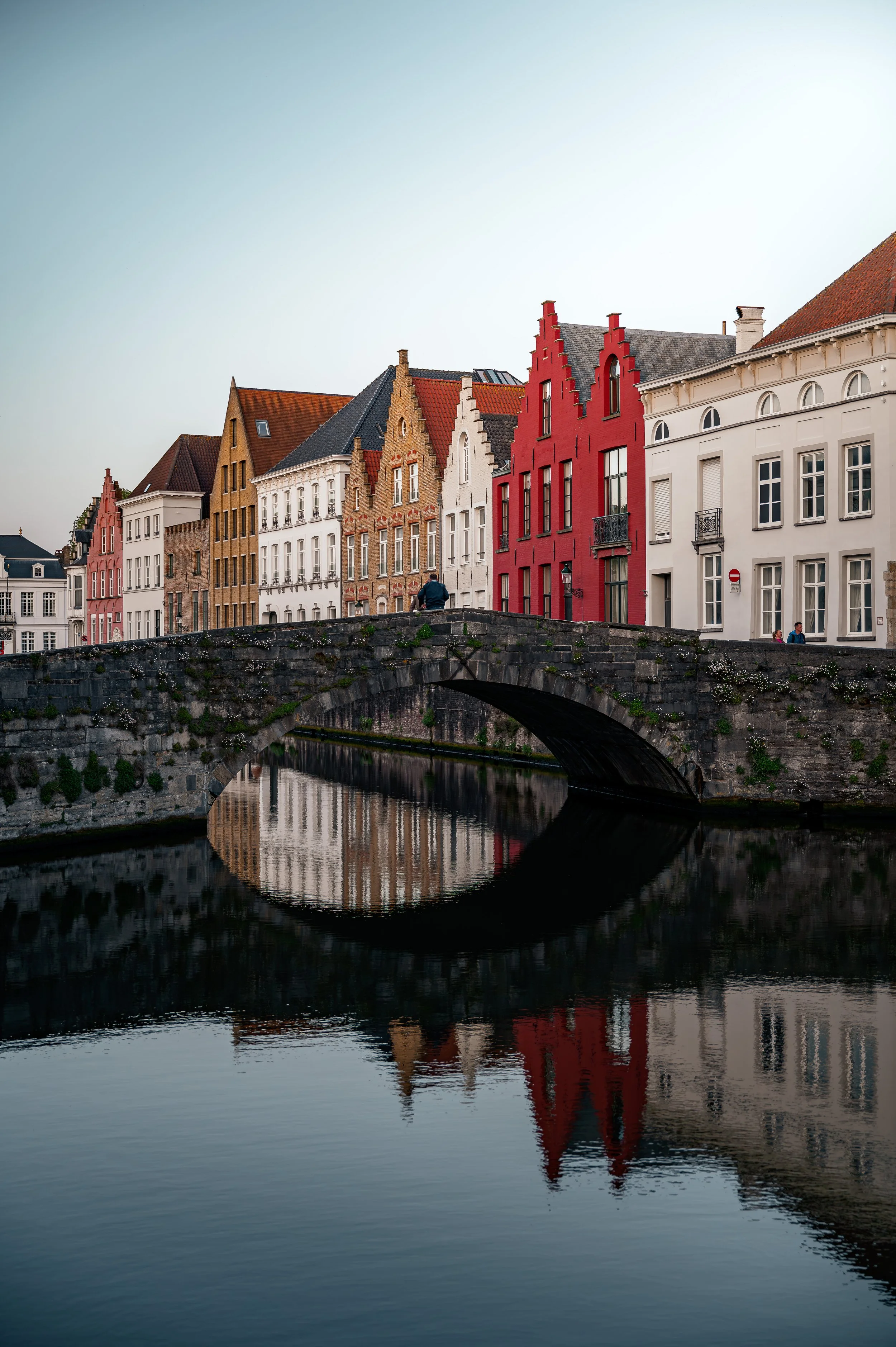 Colorful row of historic European buildings with gabled roofs along a canal, reflection in the water, stone bridge in foreground, clear sky.