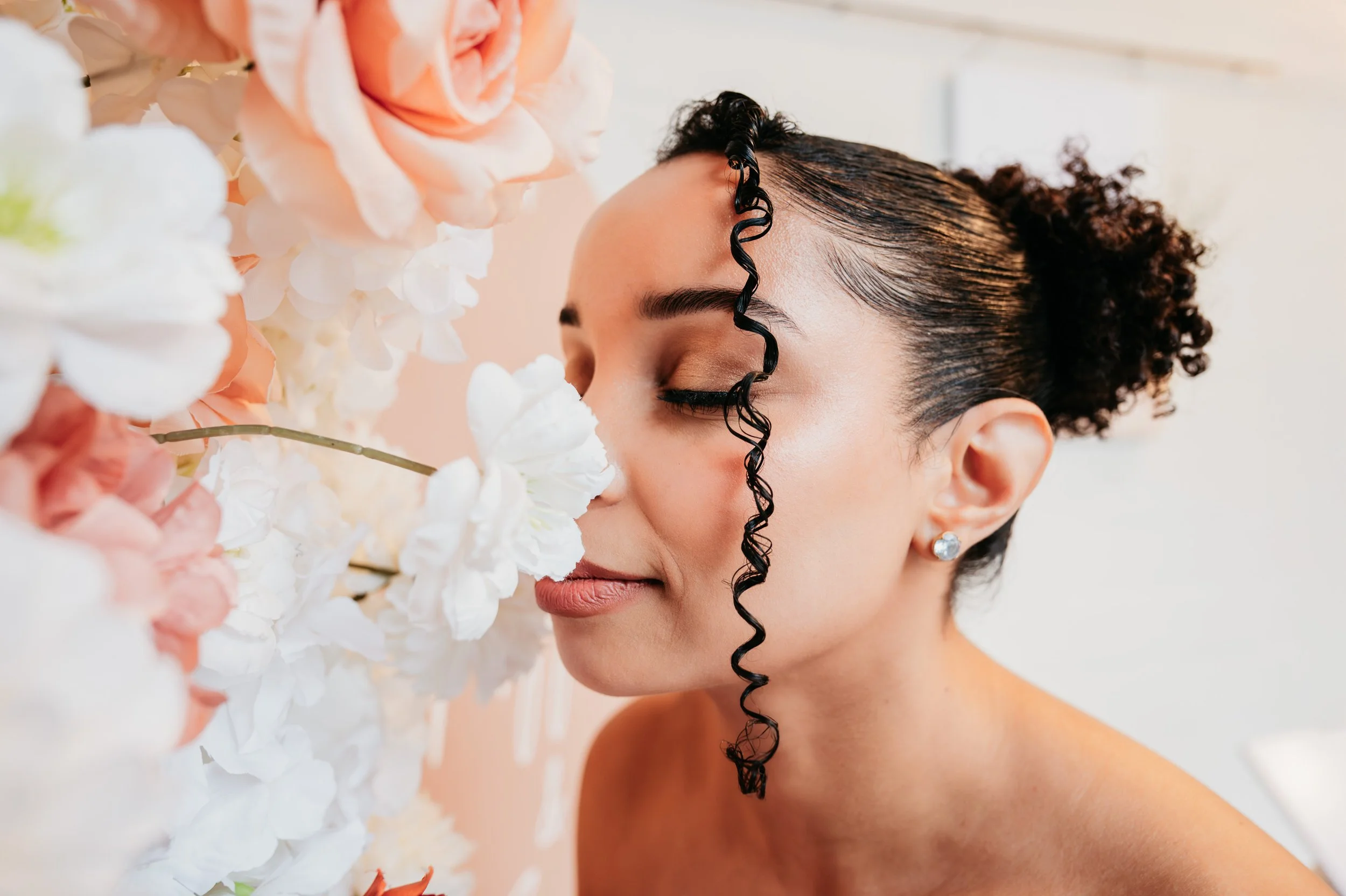 A woman with curly hair styled in a bun, wearing diamond earrings, smelling white and peach flowers.