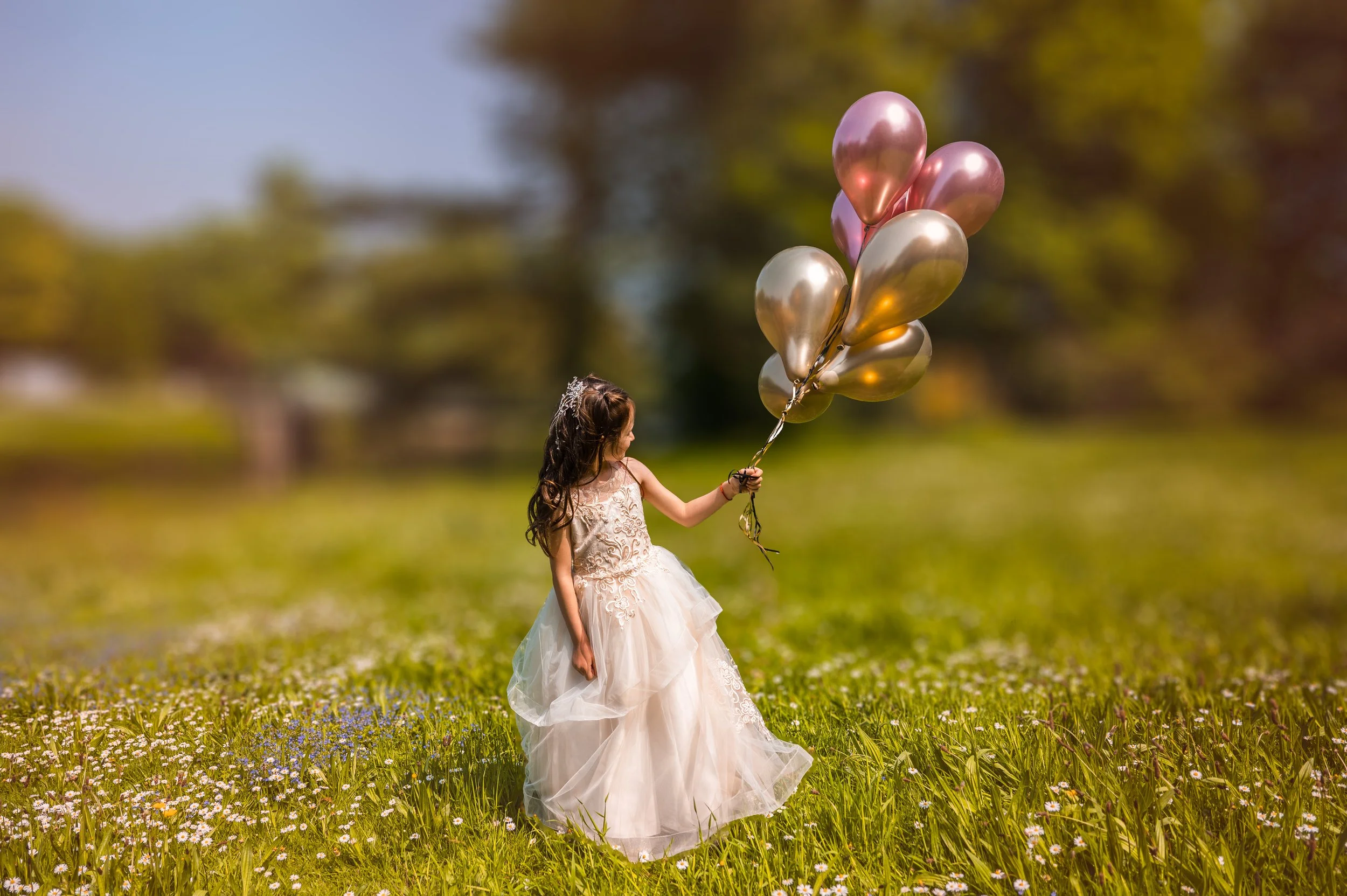 A young girl in a white lace dress holding a bunch of pink and gold balloons in a grassy field with wildflowers, trees, and blue sky in the background.