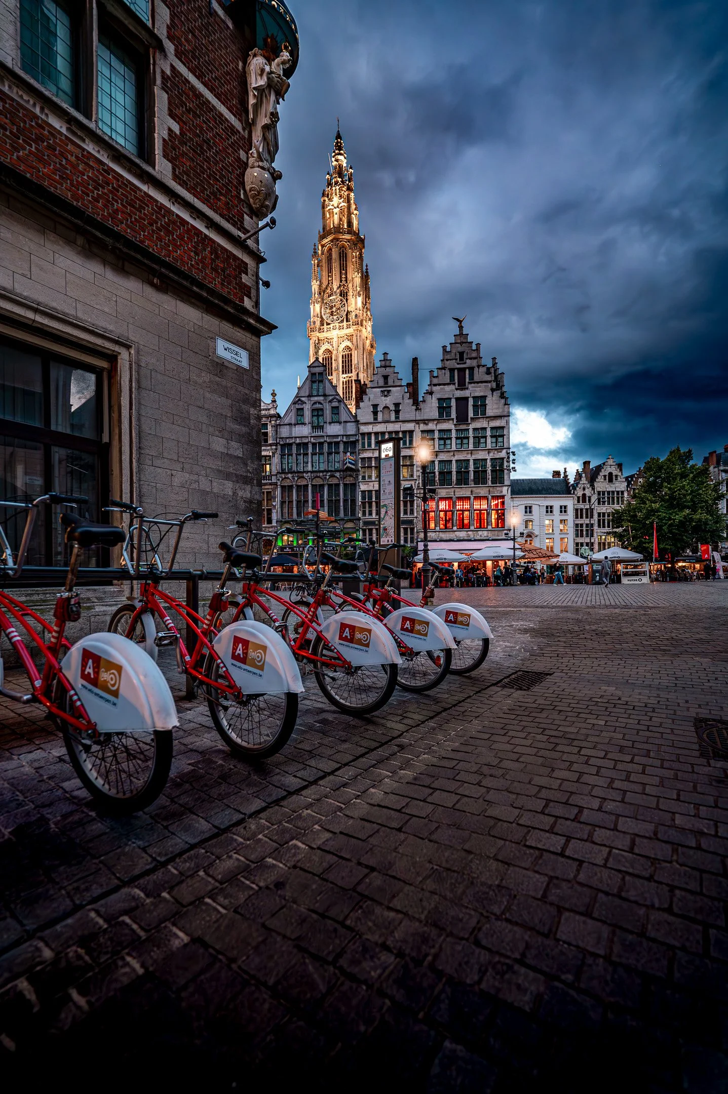Bicycle rentals parked on a brick street in front of a historic building with a tall illuminated clock tower and dark stormy skies in the background.