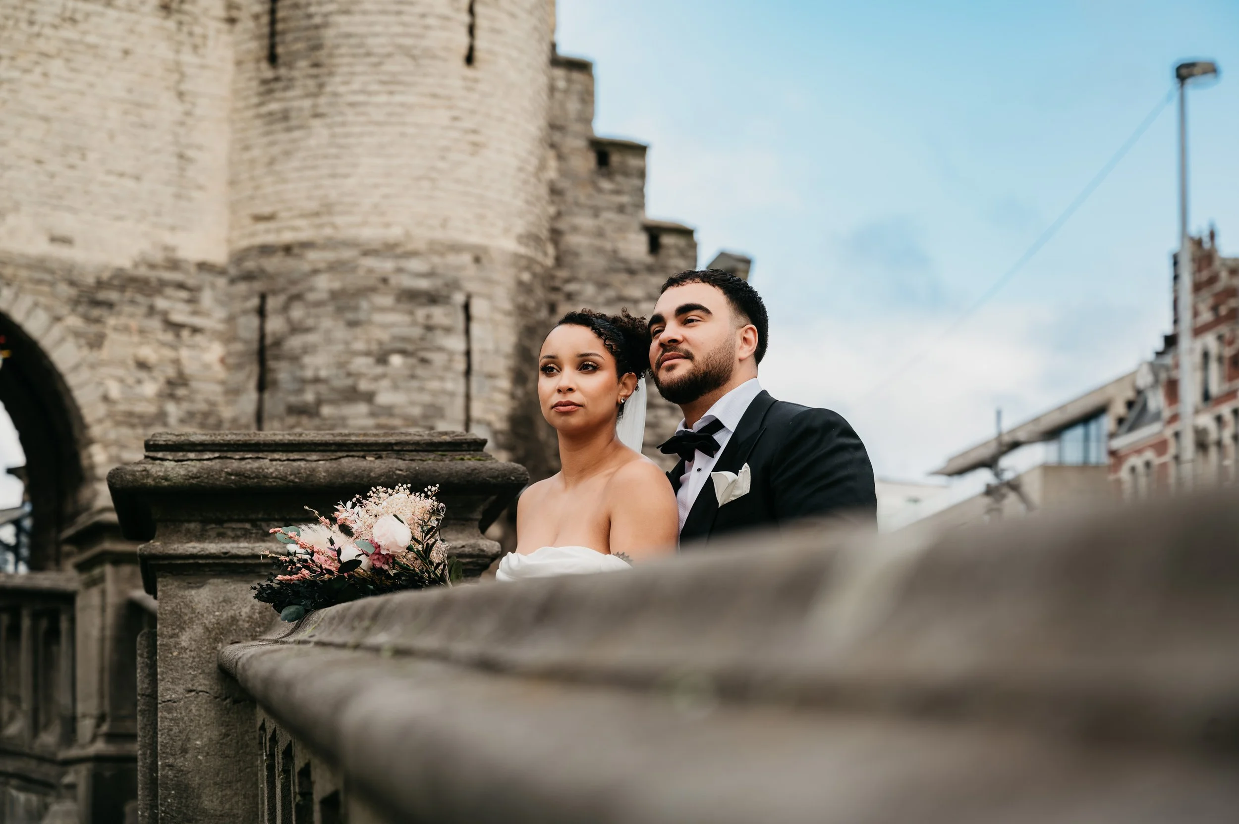 A bride and groom in wedding attire standing outdoors against a historic stone building with a tower, during daytime.