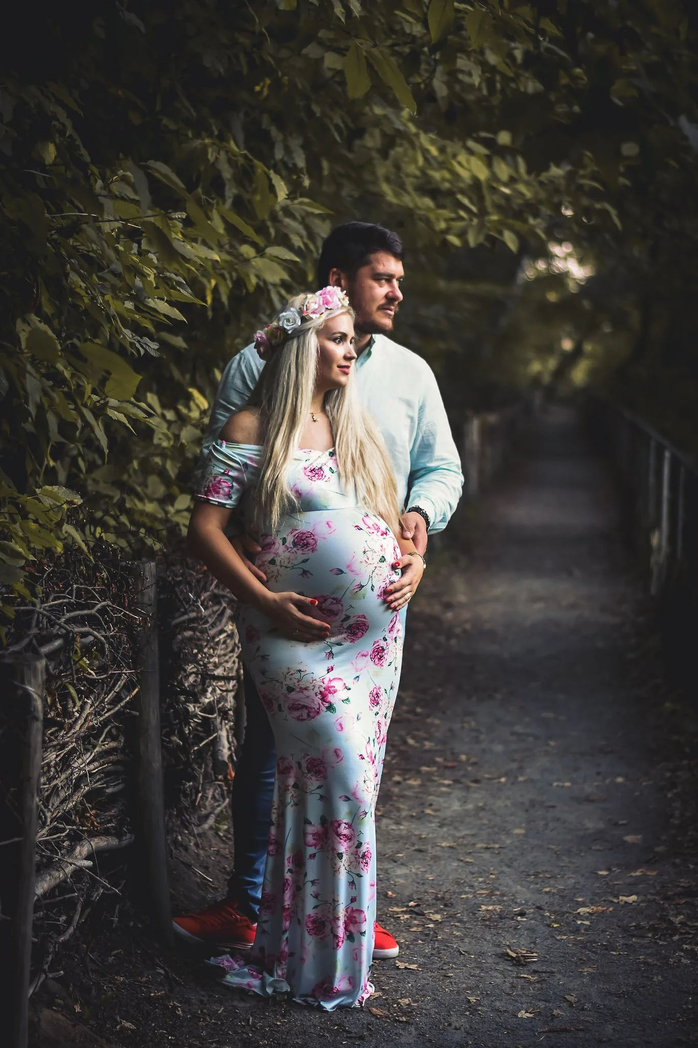 A pregnant woman with long blonde hair wearing a floral off-shoulder dress and a flower crown, standing beside a man in an outdoor setting with trees and a path, both gazing into the distance.