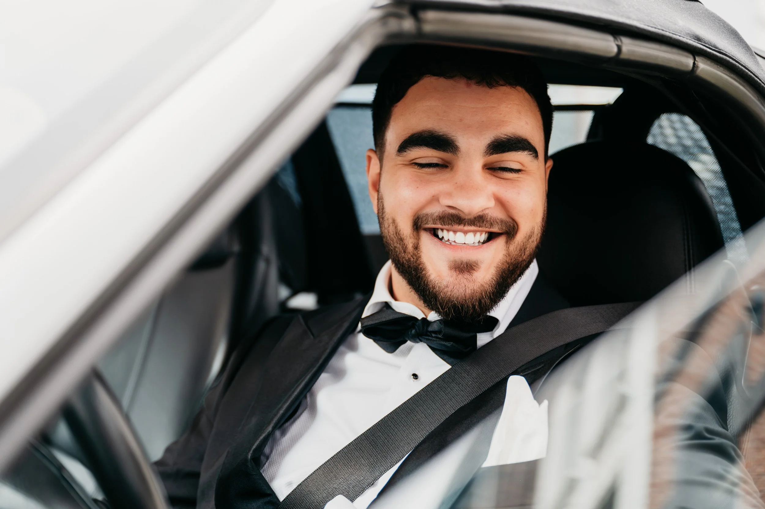 A man dressed in a tuxedo with a bow tie sitting in the driver's seat of a car, smiling happily.