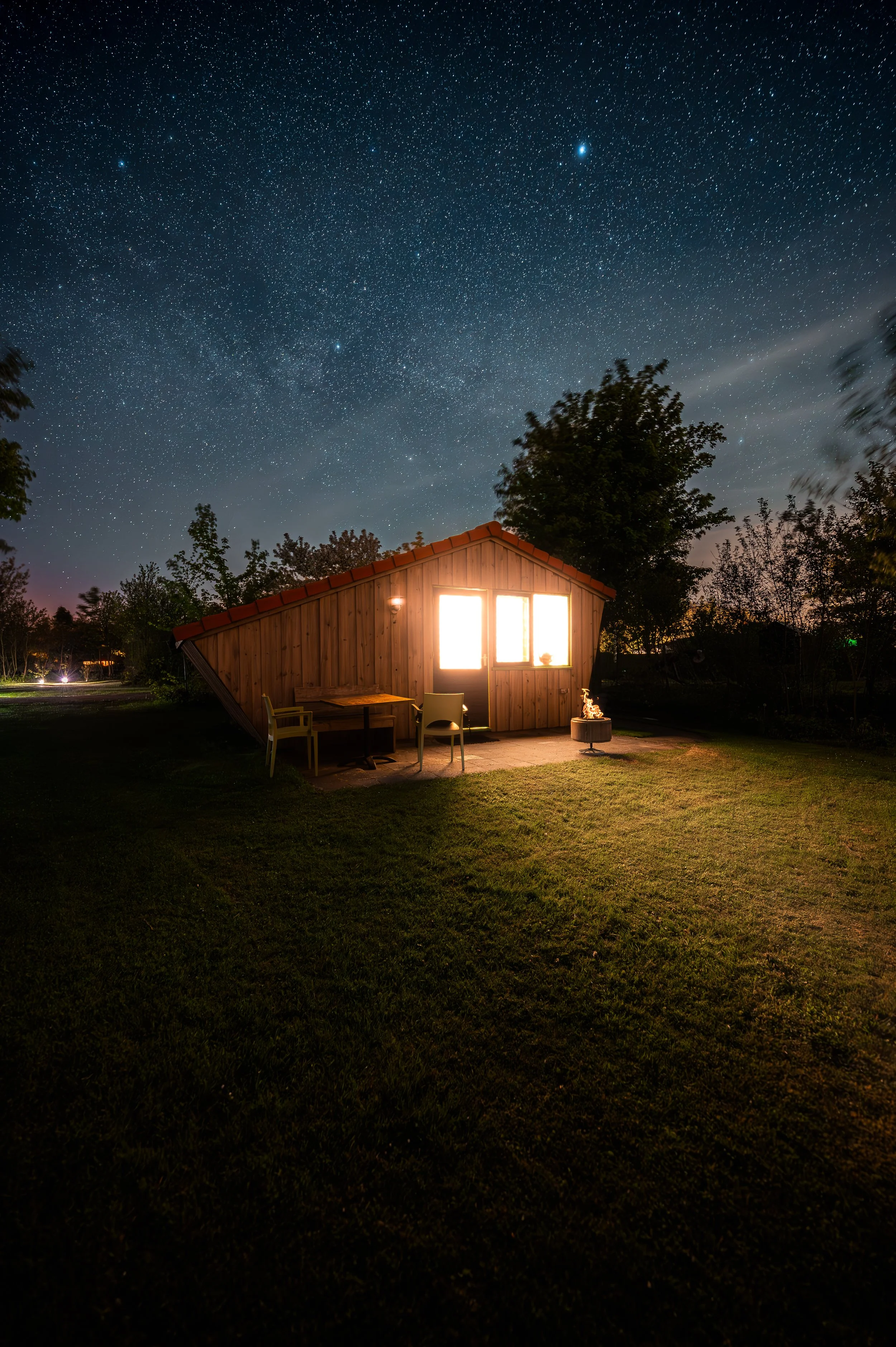 A small wooden house with bright lights on under a starry night sky, surrounded by trees and a grassy yard, with outdoor furniture and a small fire pit.