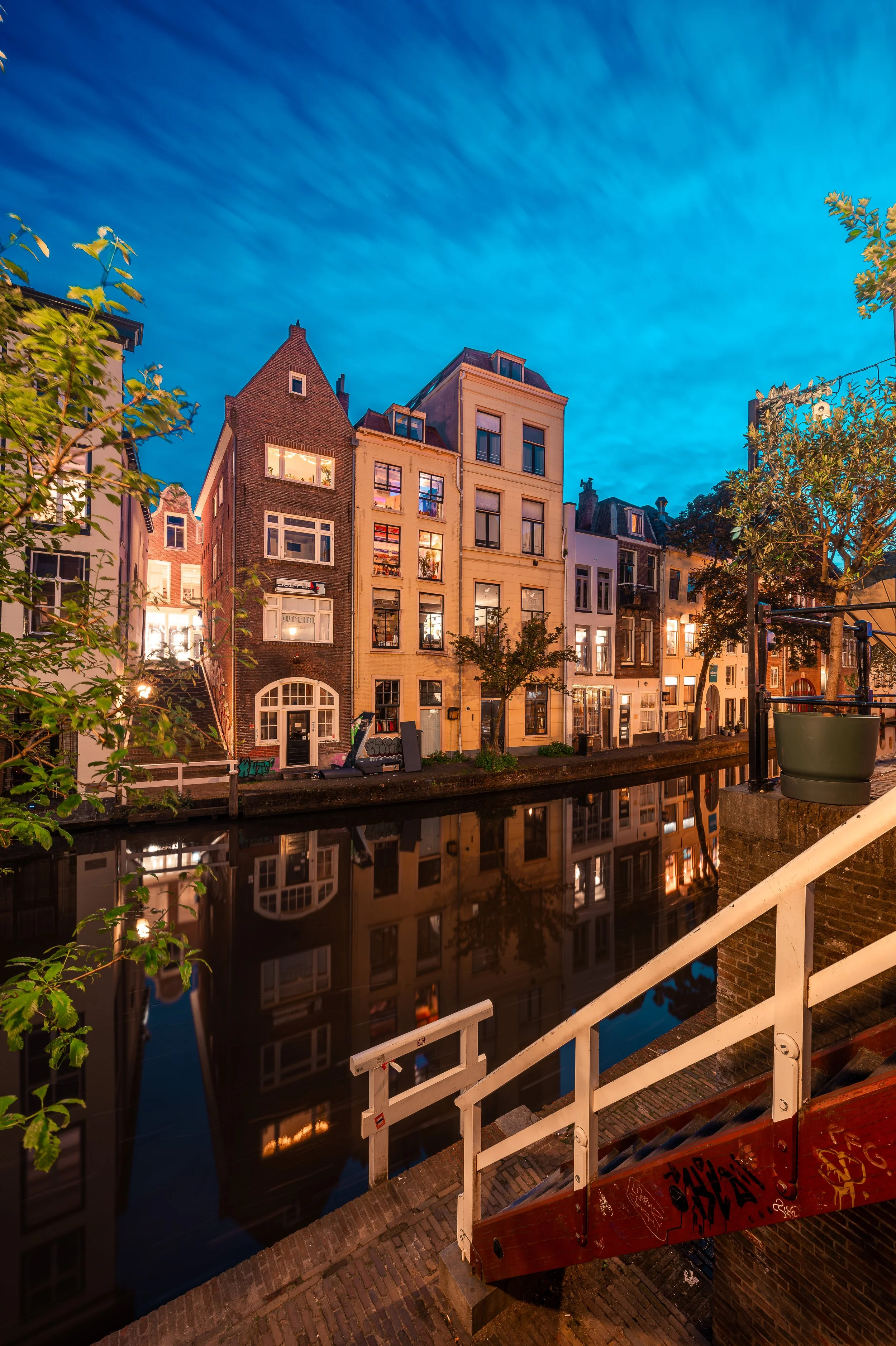 City street along a canal during twilight with illuminated buildings and their reflections in the water, trees lining the canal, and a staircase in the foreground.