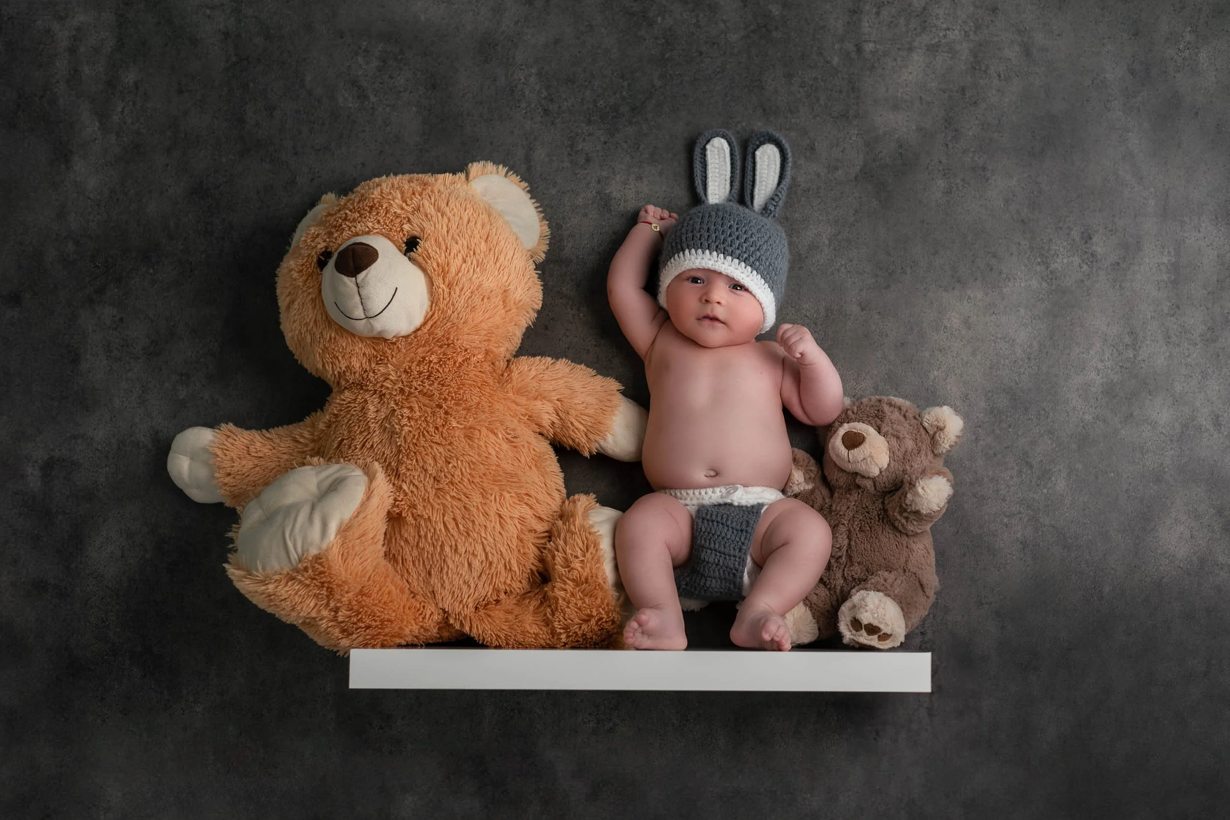 Baby lying on dark gray background with two teddy bears, one large orange plush bear on the left and a smaller brown plush bear on the right. The baby wears a gray knit hat with bunny ears, a diaper, and has one arm raised.