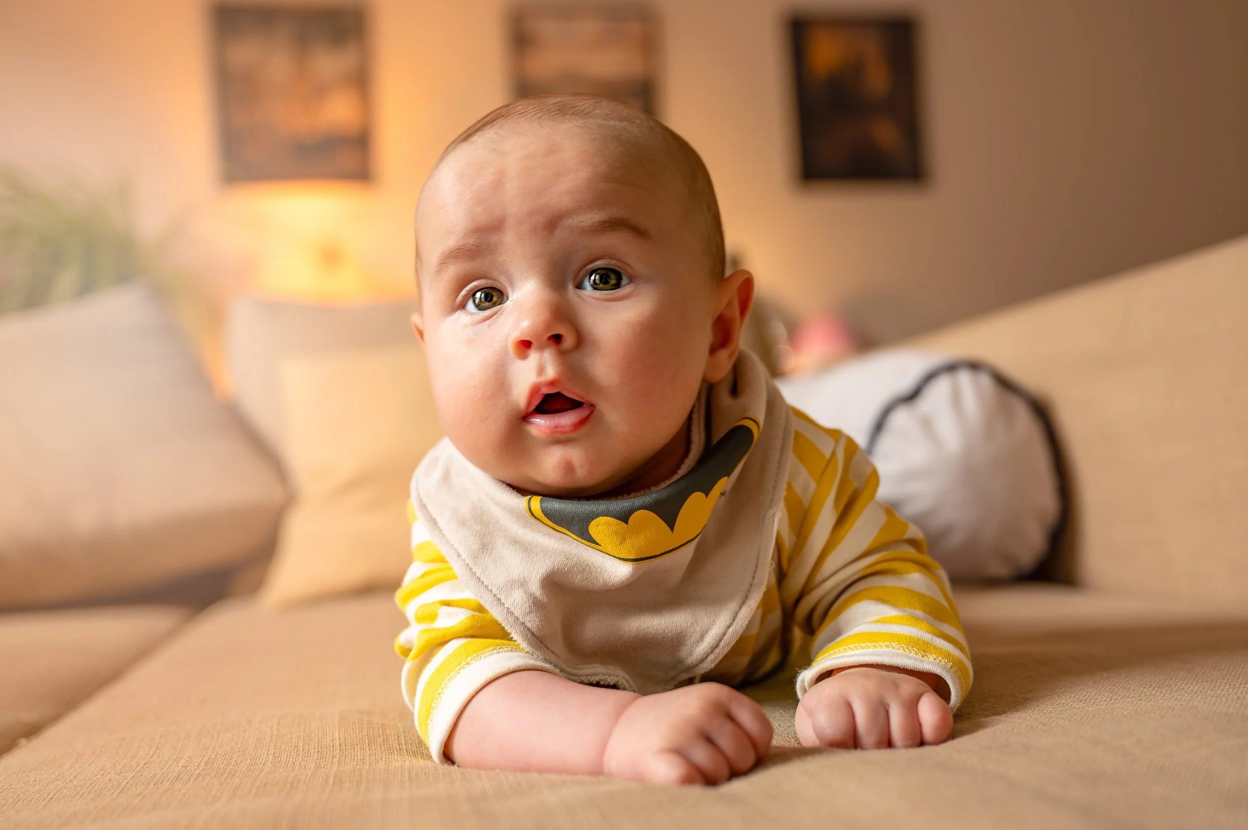 A baby crawling on a beige couch in a living room with artwork on the wall and warm lighting.