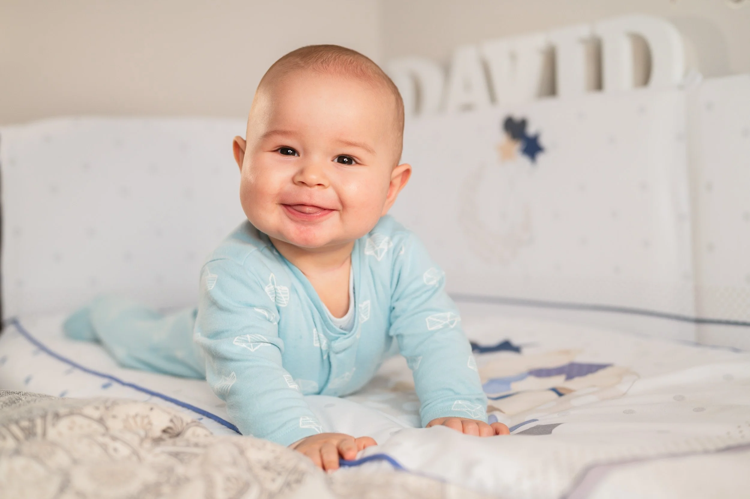 Smiling baby on a bed with white and blue bedding, wearing a light blue onesie with white patterns, with a blurred white headboard in the background.