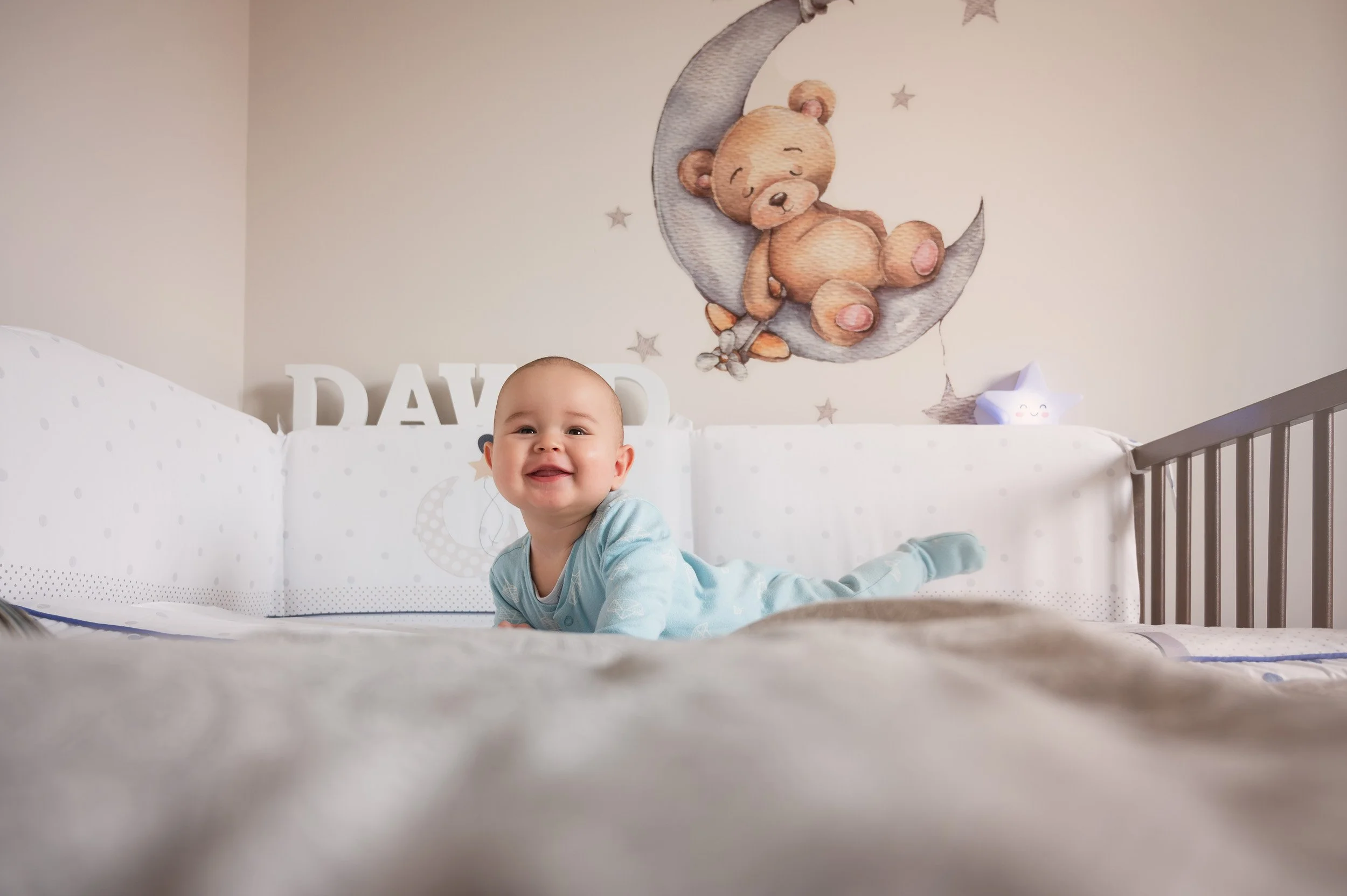A happy baby lying on its stomach in a crib, smiling at the camera, with a mural of a teddy bear sitting on a crescent moon behind him.