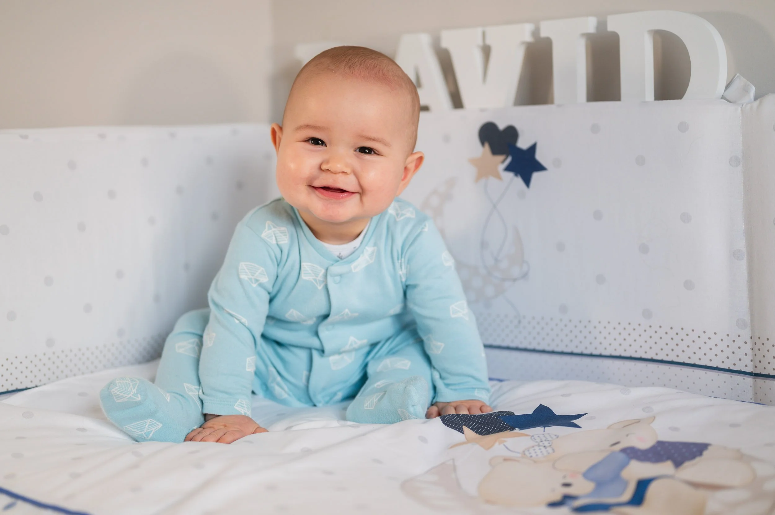 Smiling baby in blue pajamas sitting on a crib mattress with decorative stars and a bear illustration.