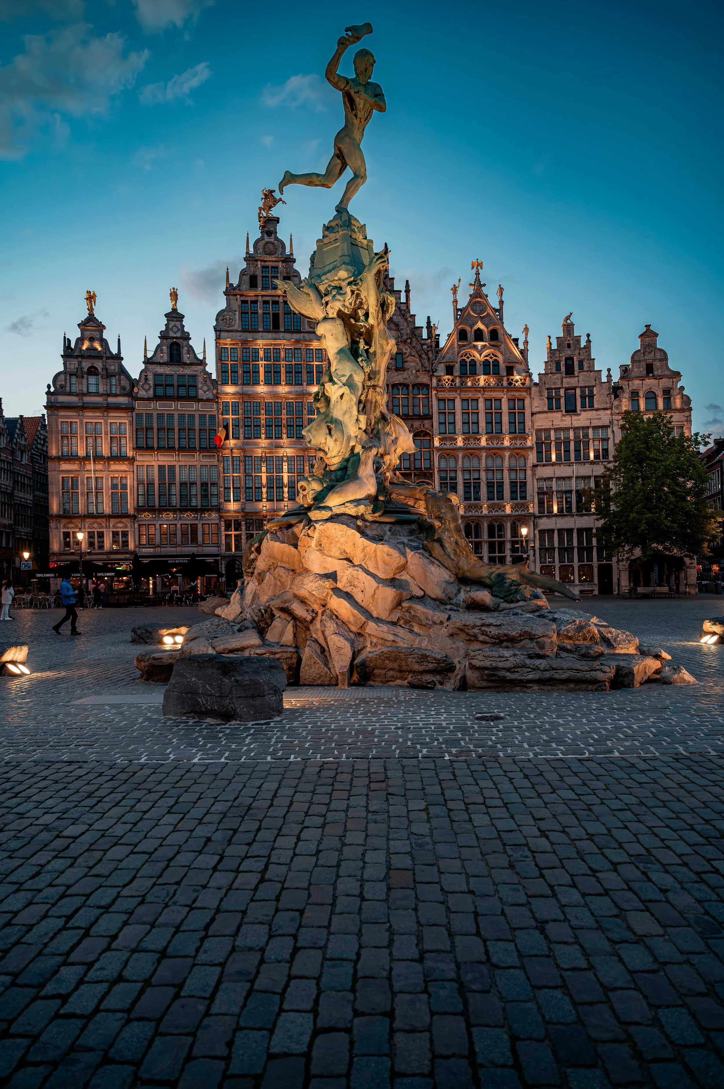 A statue of a boy holding a fish stands tall in a European city square at dusk, with historic buildings in the background and cobblestone pavement in the foreground.