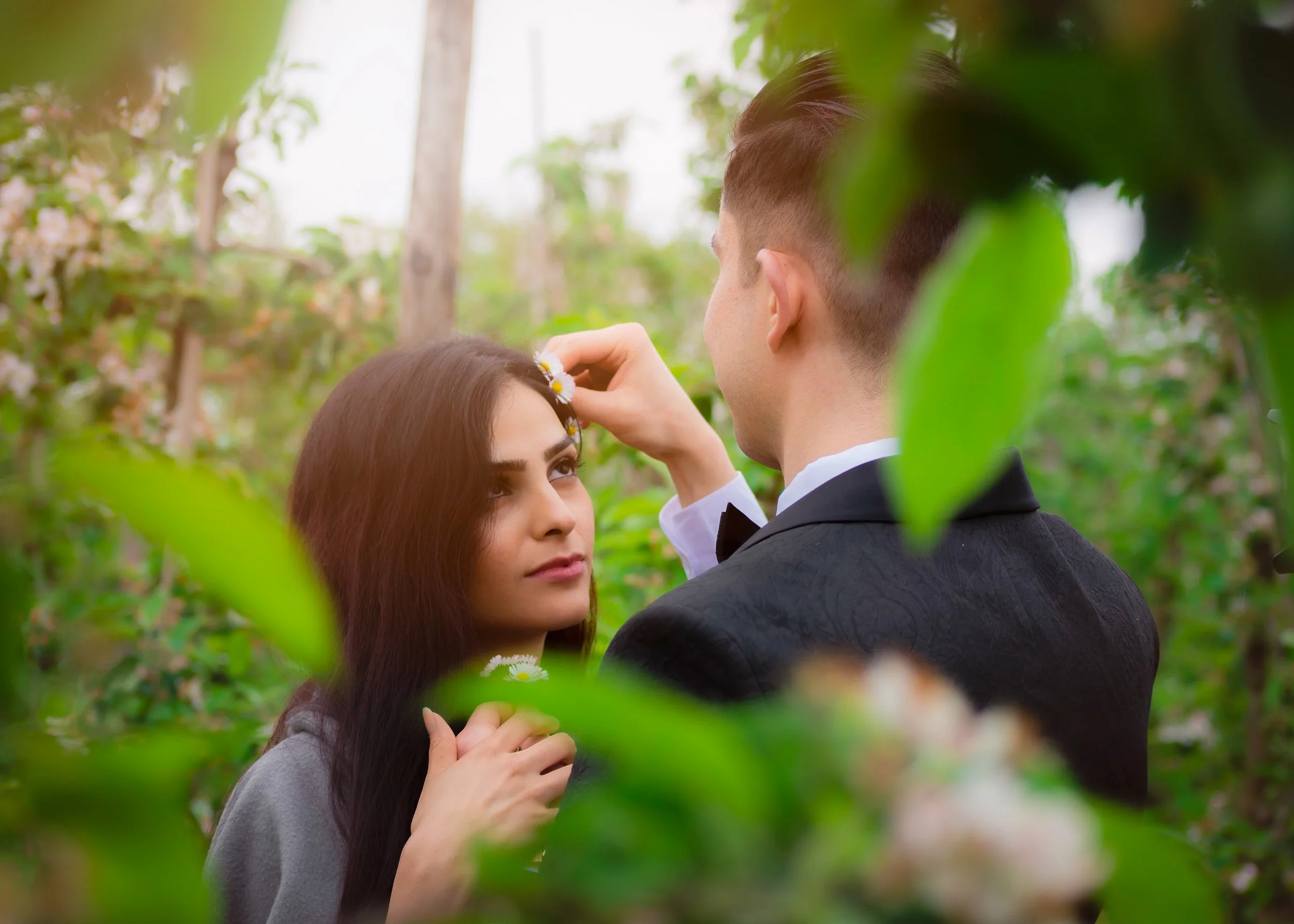 A young couple in formal attire, surrounded by greenery, with the man touching a flower near the woman's head.