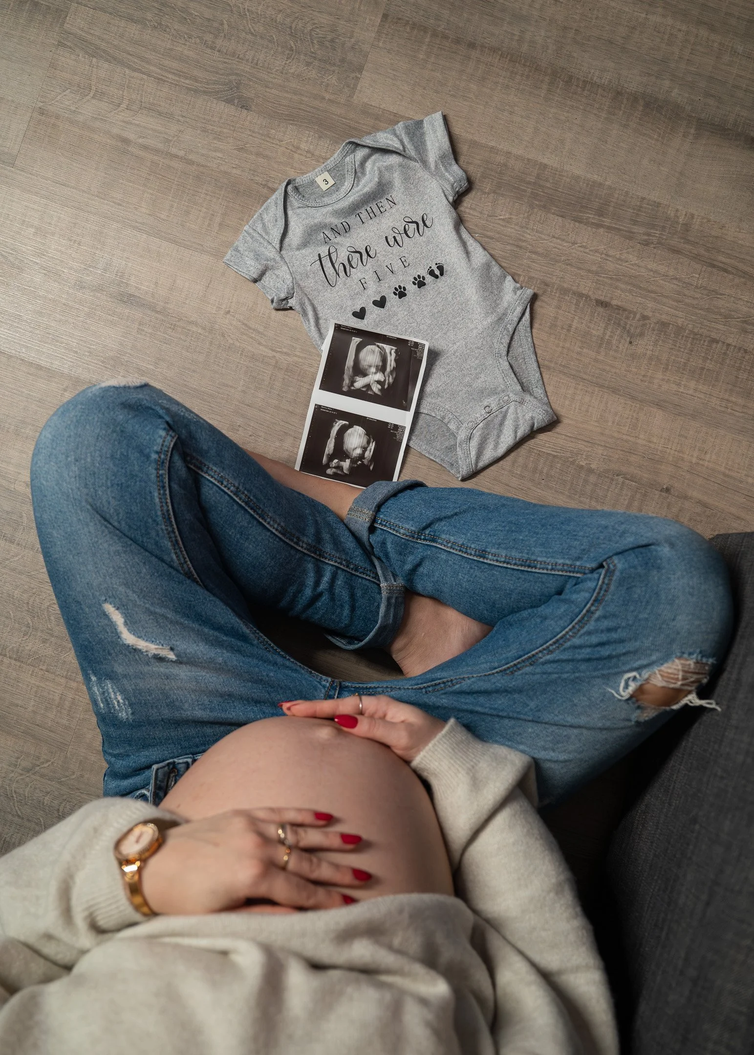 A pregnant woman sitting on a couch with her hand on her belly, wearing ripped jeans and a beige sweater. On the floor in front of her are ultrasound photos and a gray baby onesie with the words "And then there were five" and paw prints.