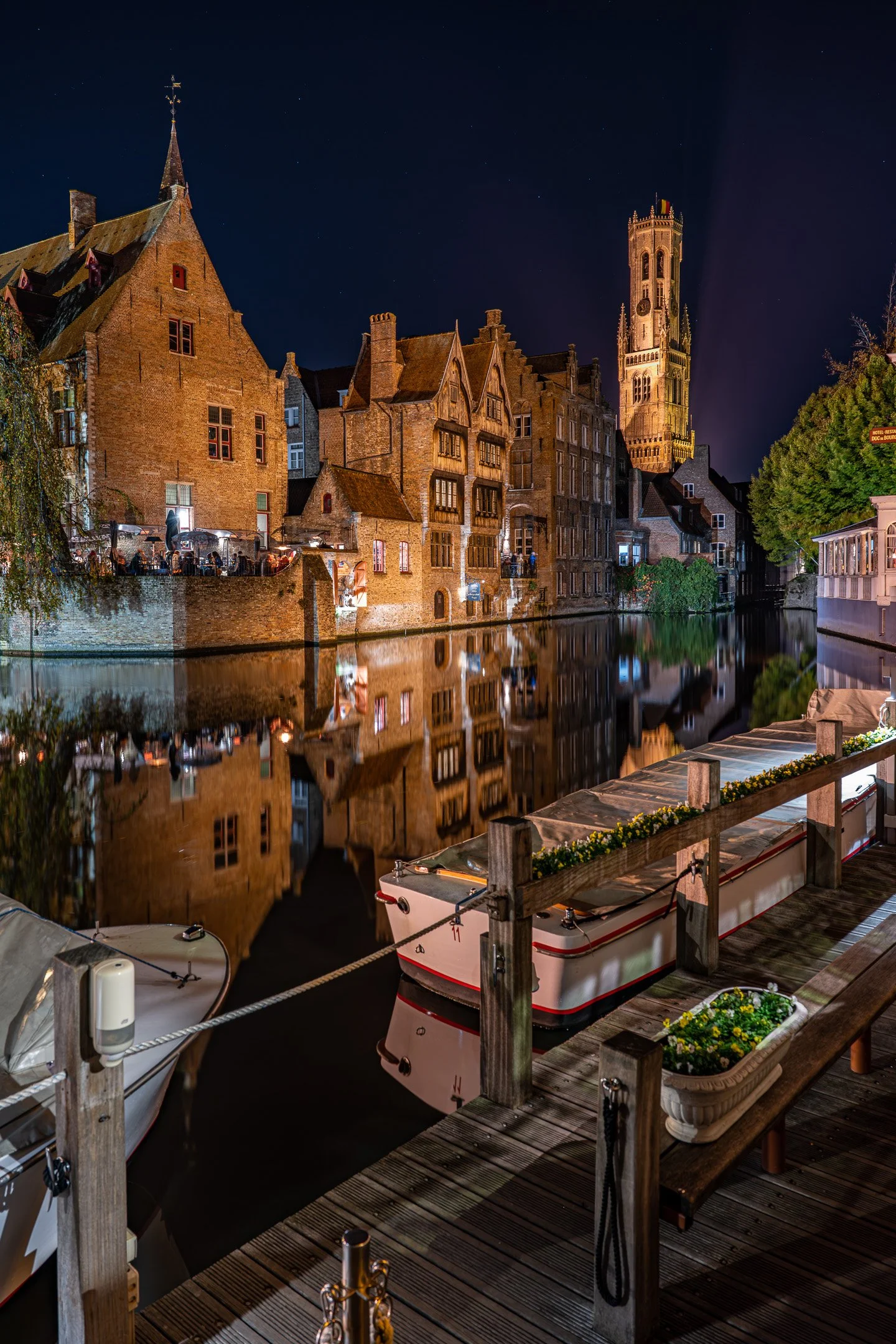 Night view of historic brick buildings along a canal, with a tall church tower in the background, illuminated and reflected in the water, with boats moored along a wooden dock in the foreground.