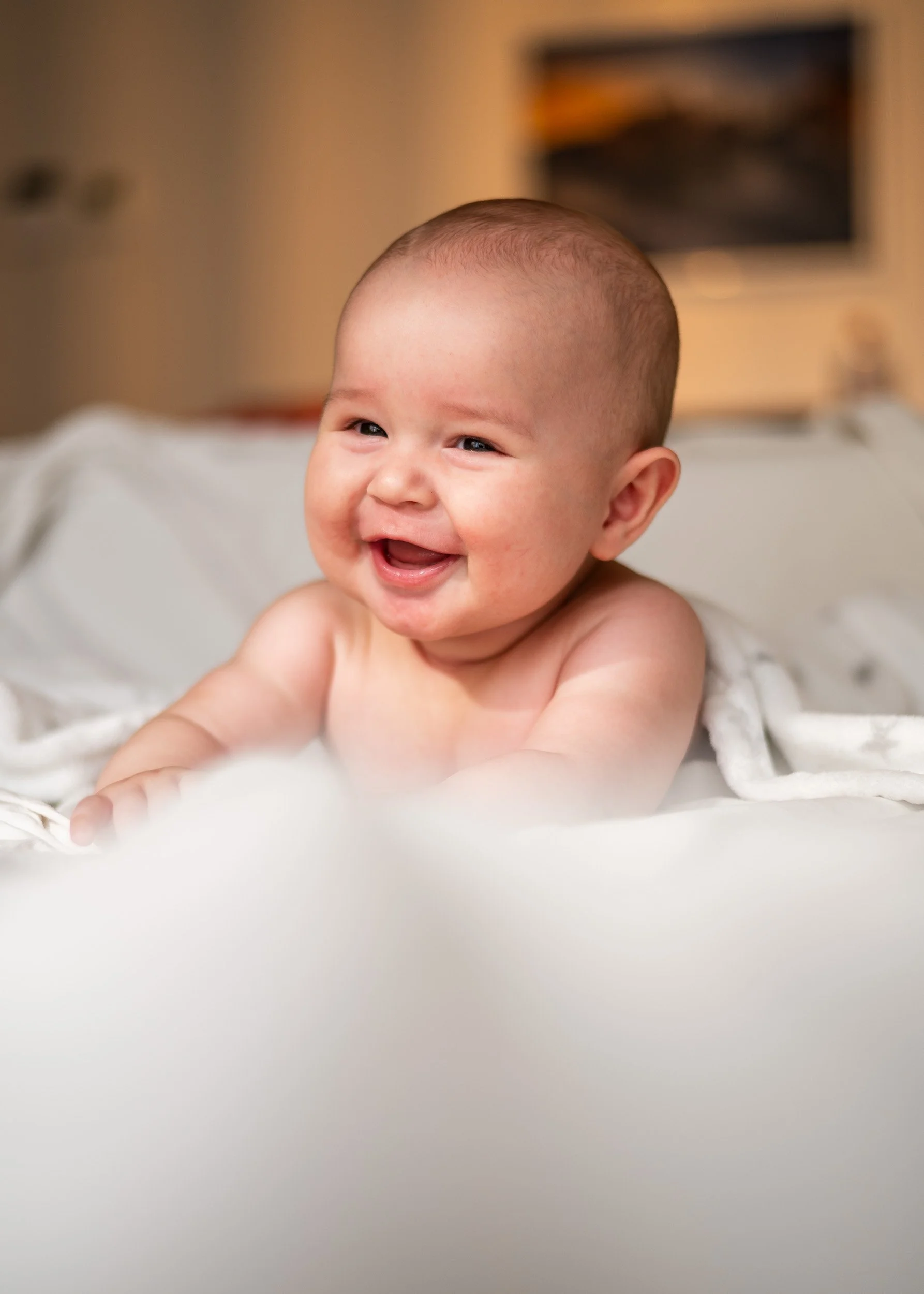 A smiling baby with no shirt, lying on a bed with a white blanket, in a warmly lit room.