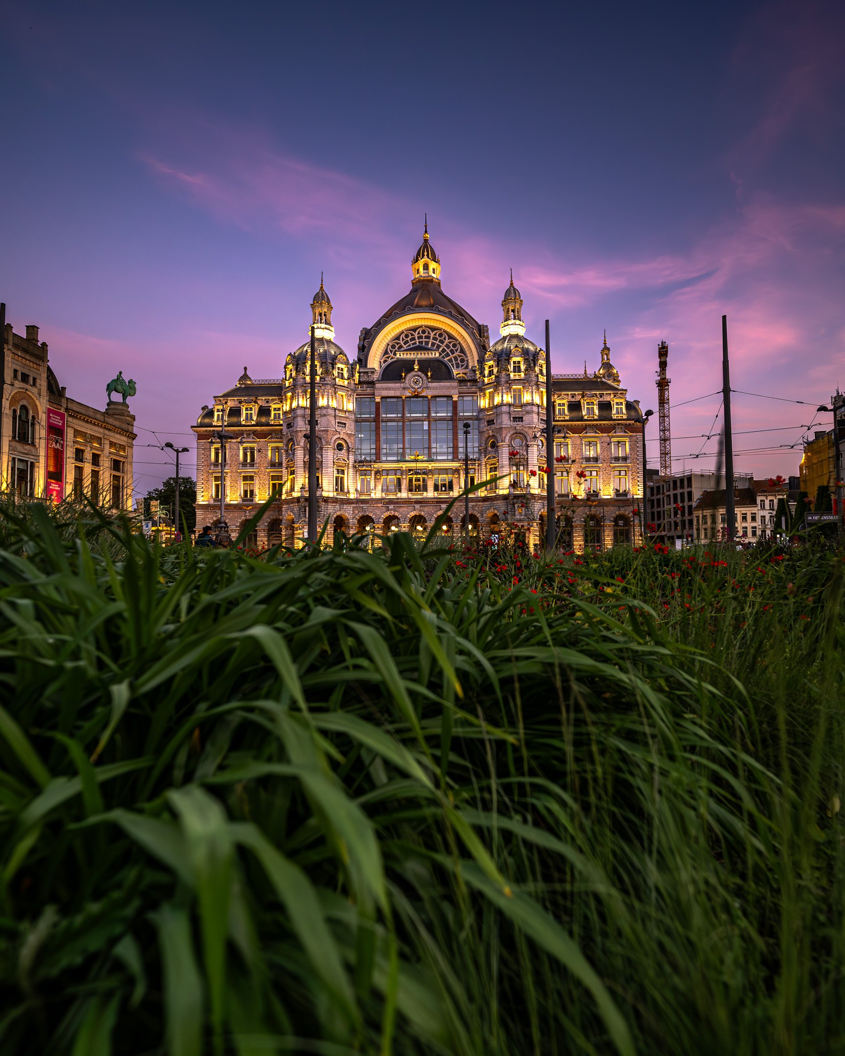 A grand, illuminated historic building with domes and turrets, seen at dusk with a purple sky, surrounded by green grass and flowers in the foreground.