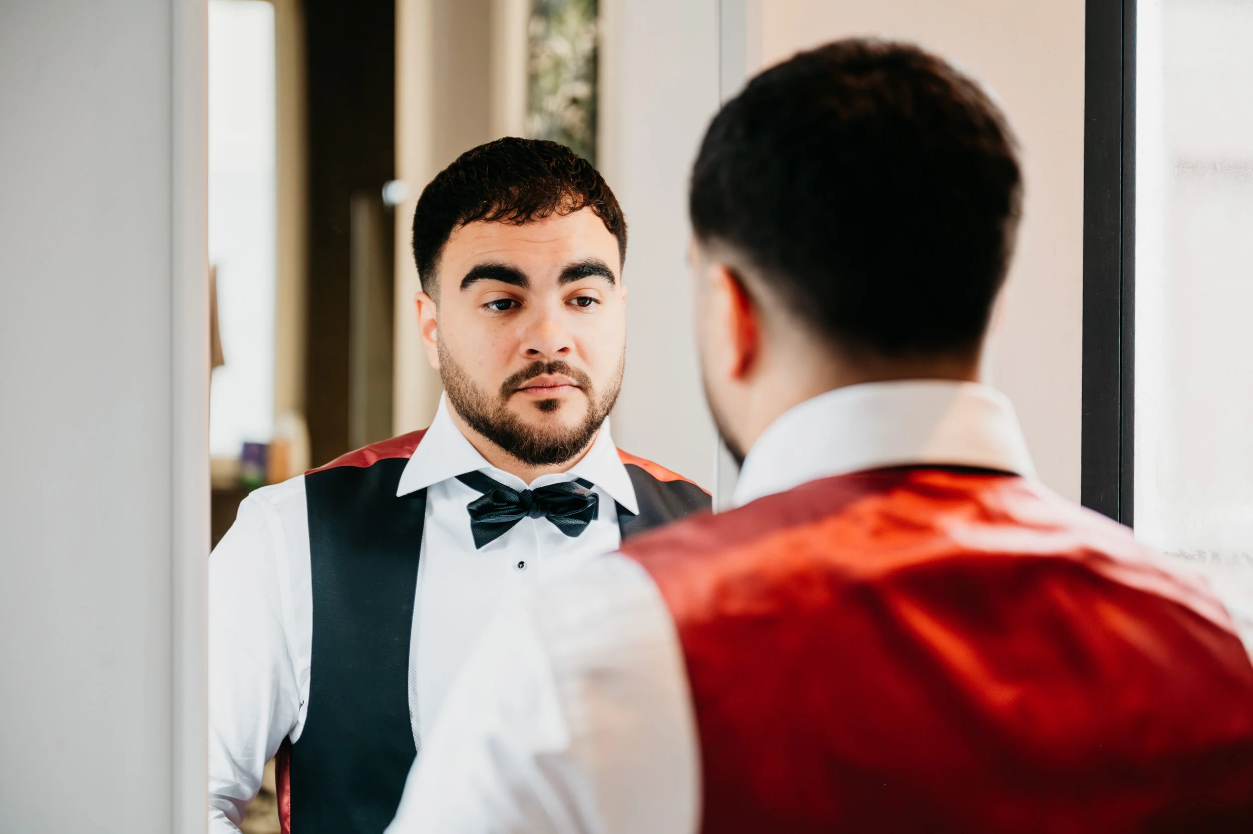A man in formal attire, including a tuxedo with a bow tie and red and black blazer, looks at his reflection in a mirror.